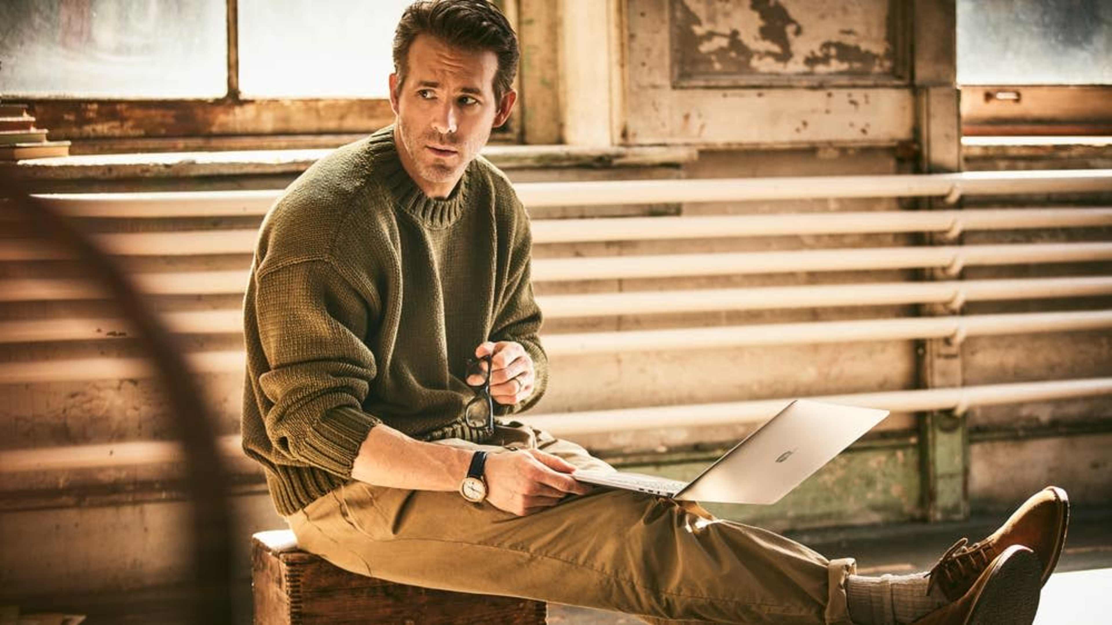 A man sits on a wooden box in a sunlit room, working on a laptop while holding a pair of glasses.