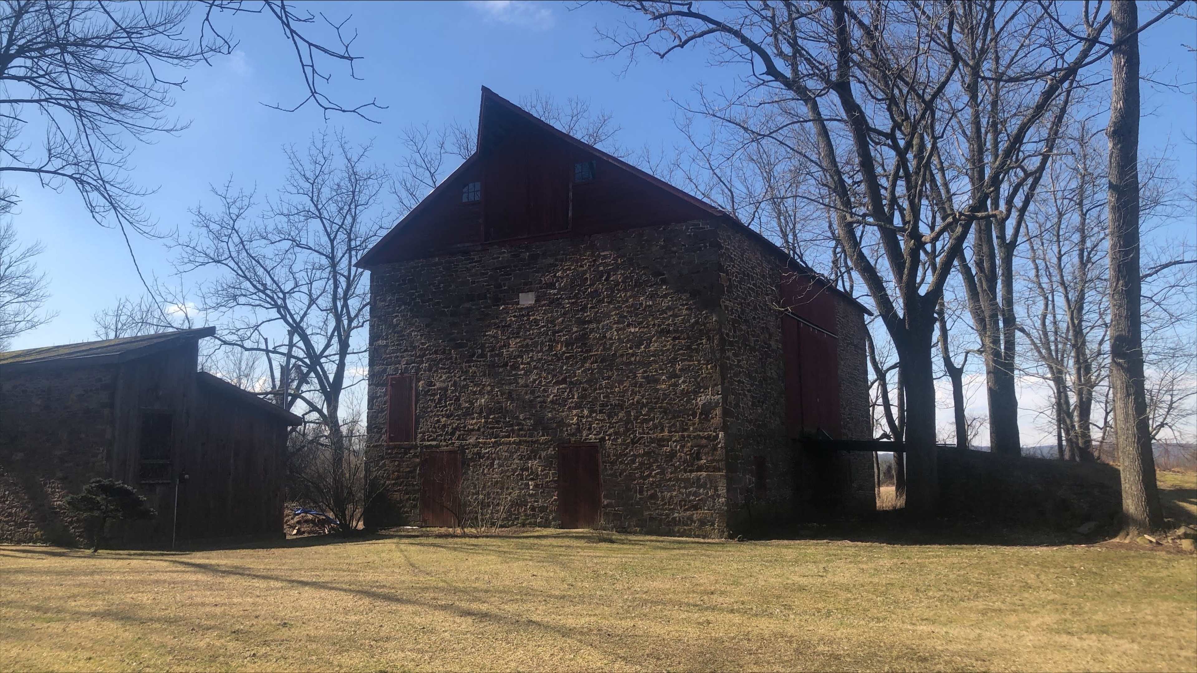 The image shows a large, stone barn with a red roof, situated in a grassy area surrounded by leafless trees.