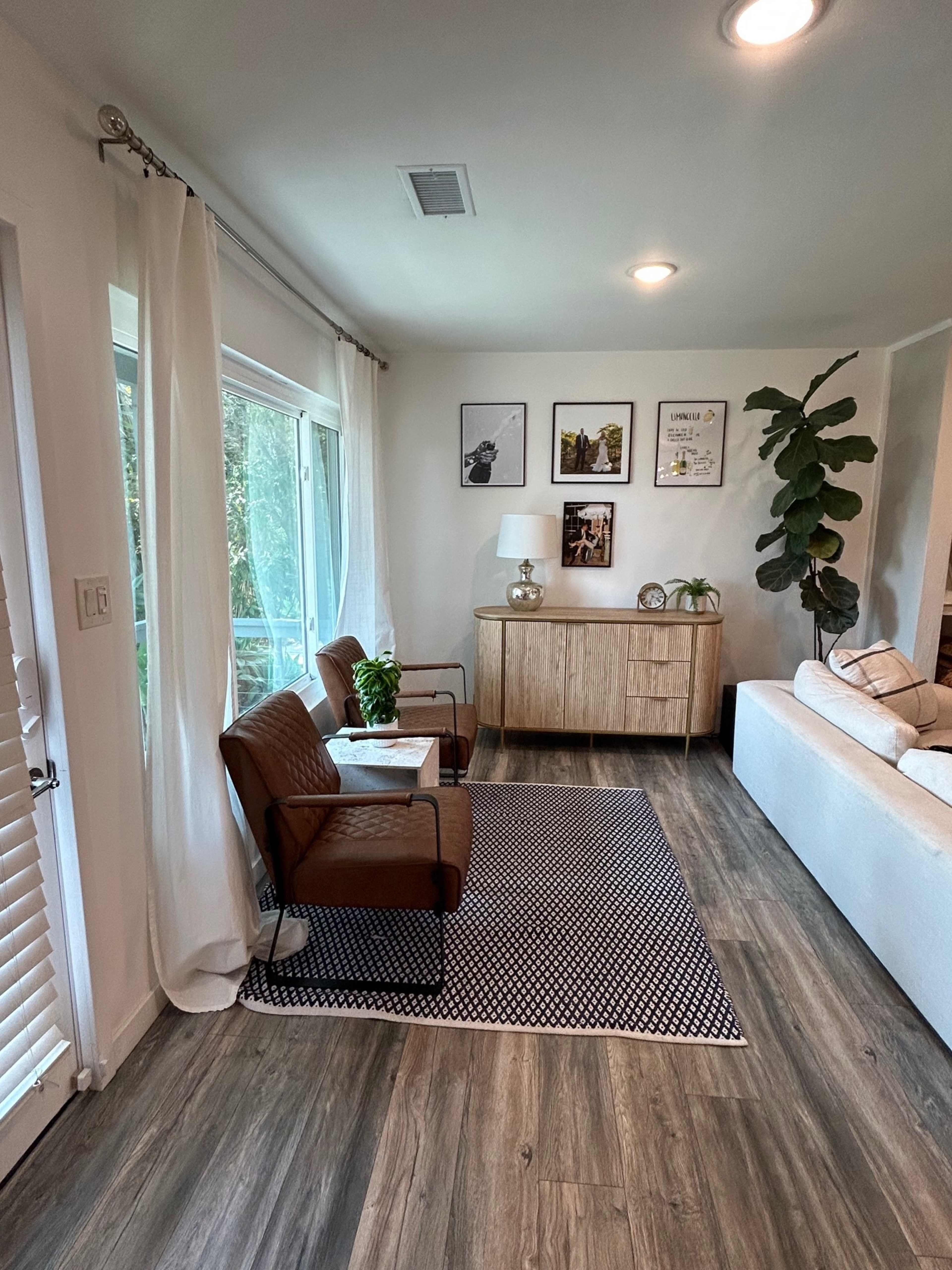 A cozy living room features two brown chairs, a modern side table, a light wood cabinet, and large windows with natural light.