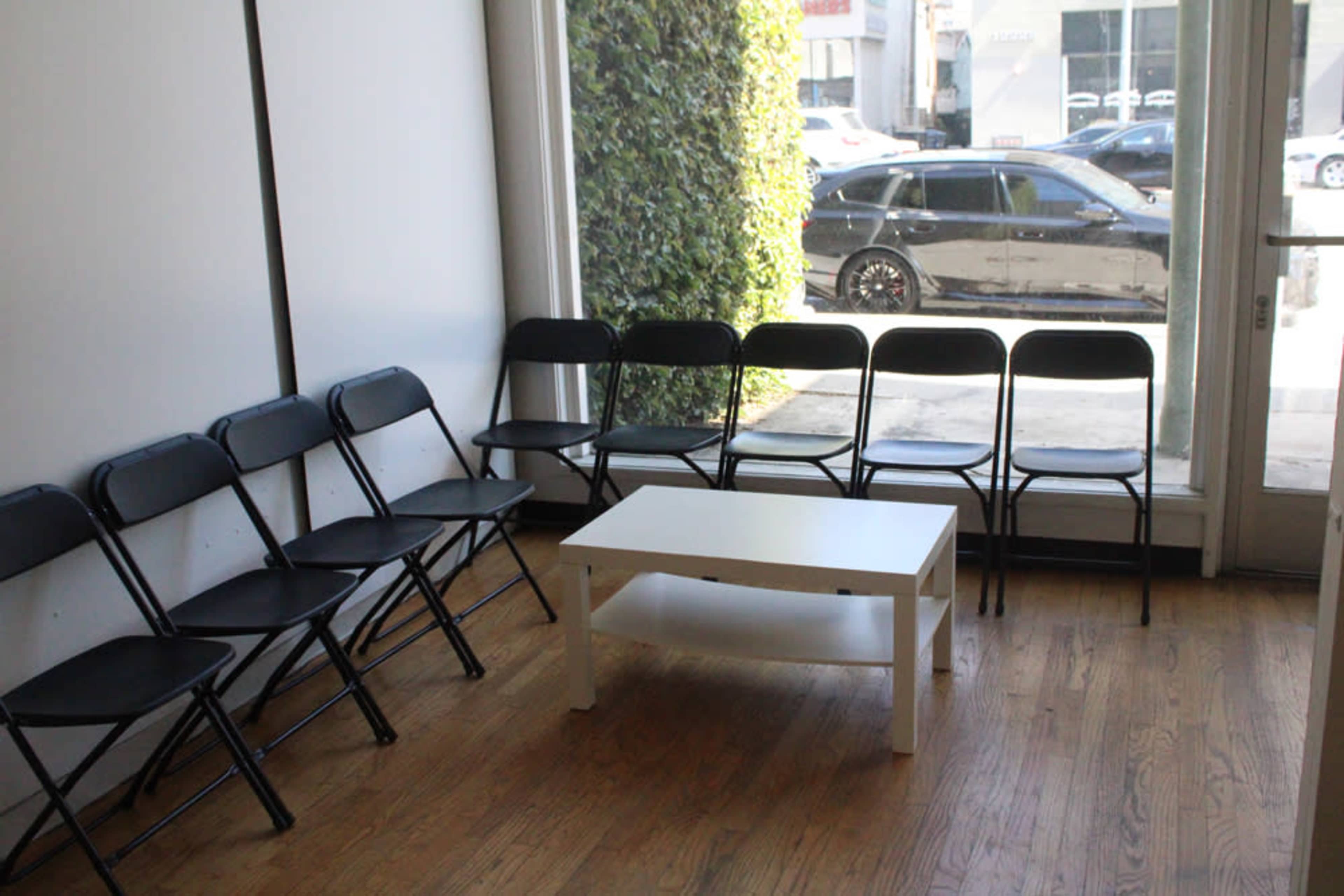 The image shows a waiting area with a row of black folding chairs and a white coffee table facing a large window that overlooks a street.