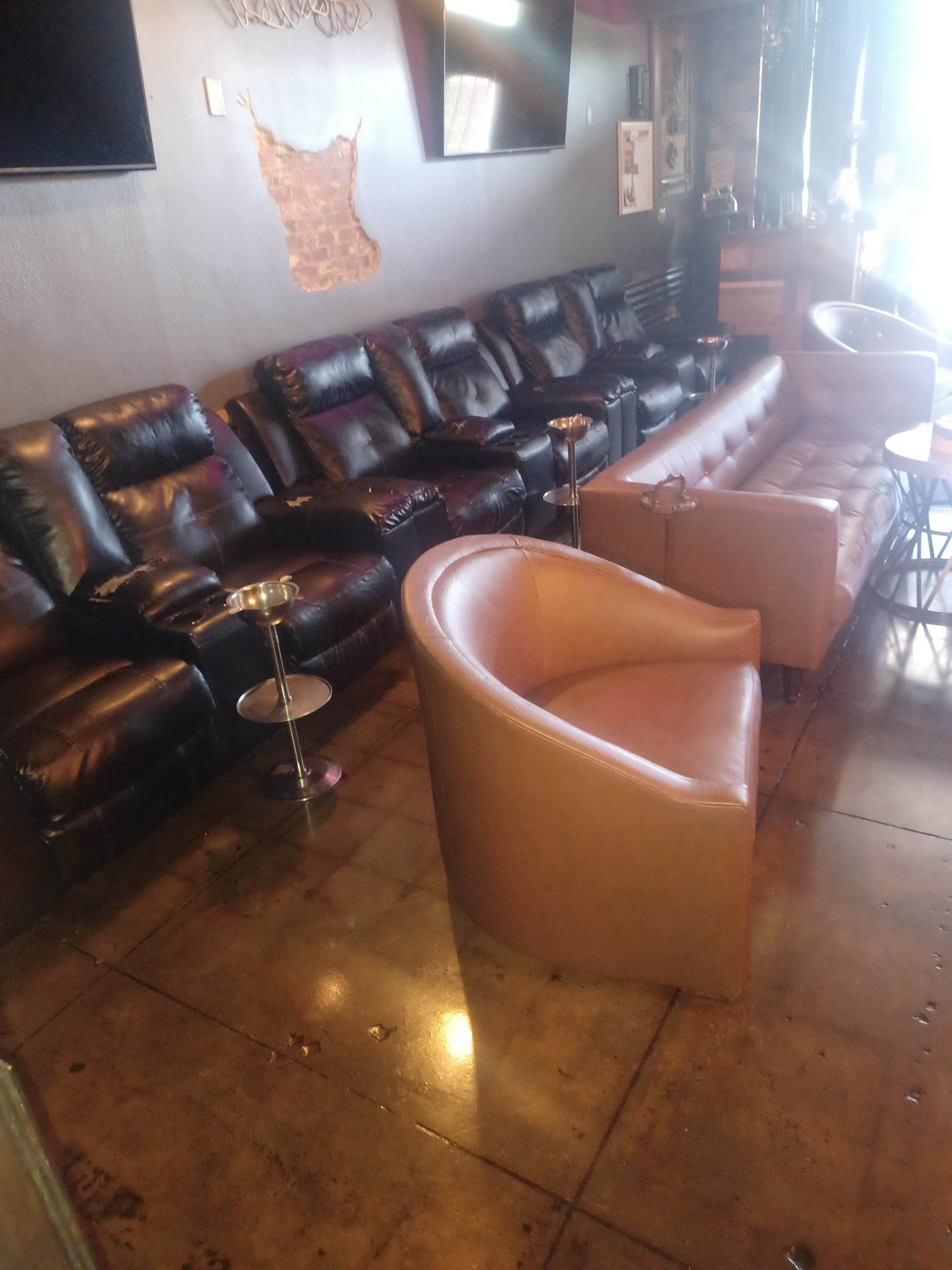A lounge area with black leather recliners, a brown upholstered sofa, and circular side tables arranged on a polished concrete floor.