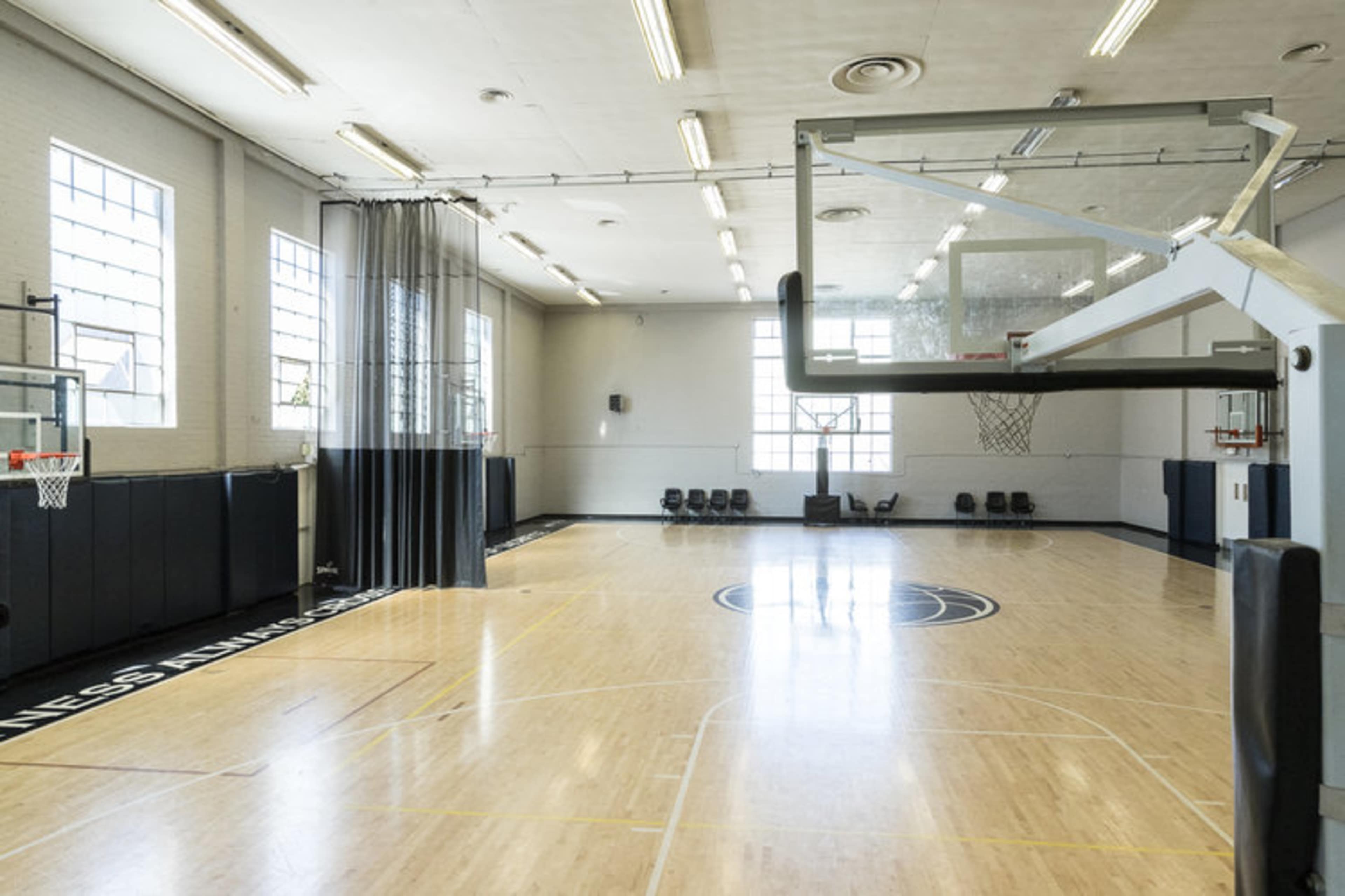 An empty basketball gymnasium with two hoops, polished wooden floors, and large windows allowing natural light to enter.