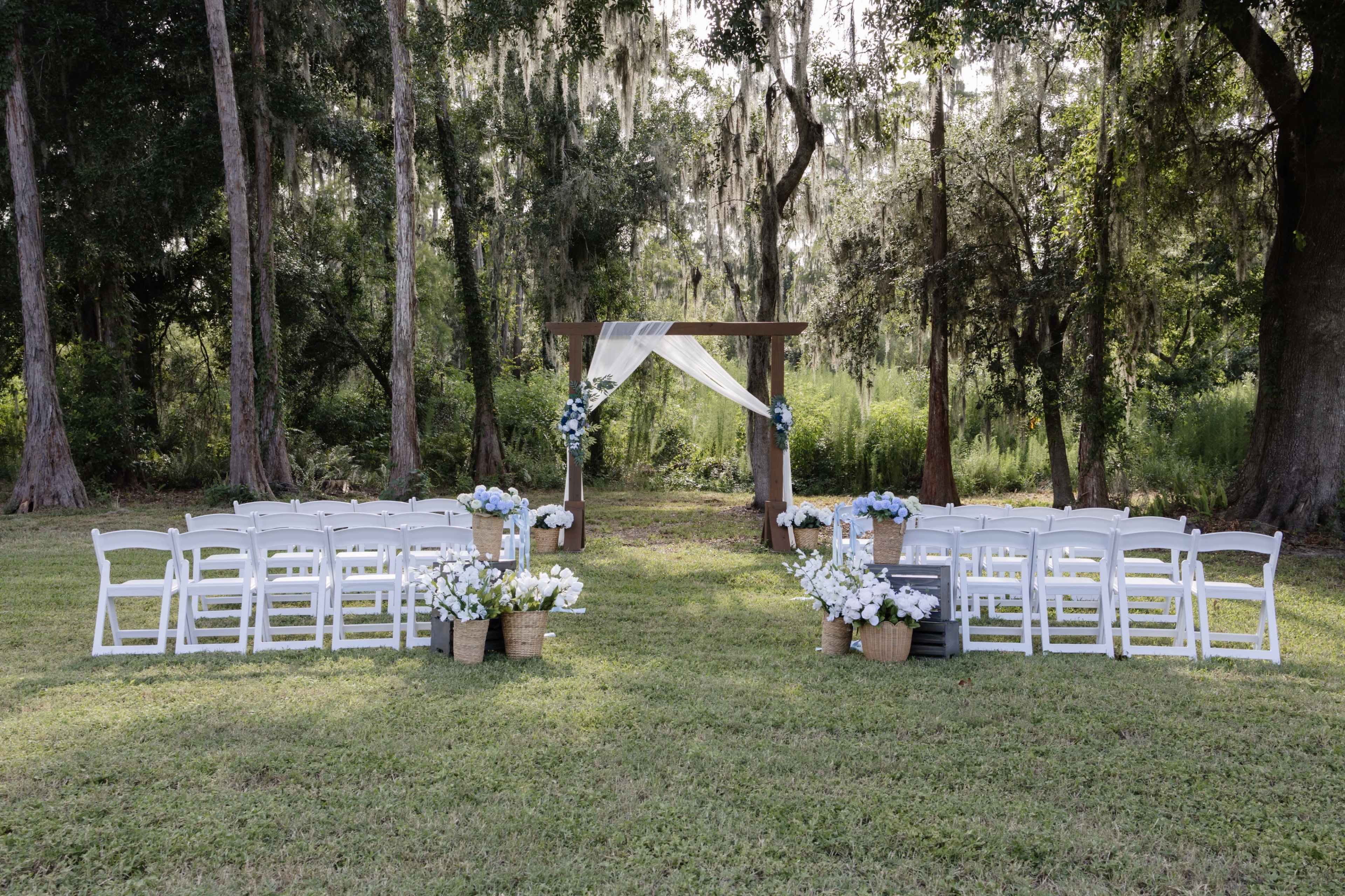 The image shows a wedding ceremony setup in a grassy area, featuring rows of white chairs facing a decorated wooden arch adorned with drapery and floral arrangements.