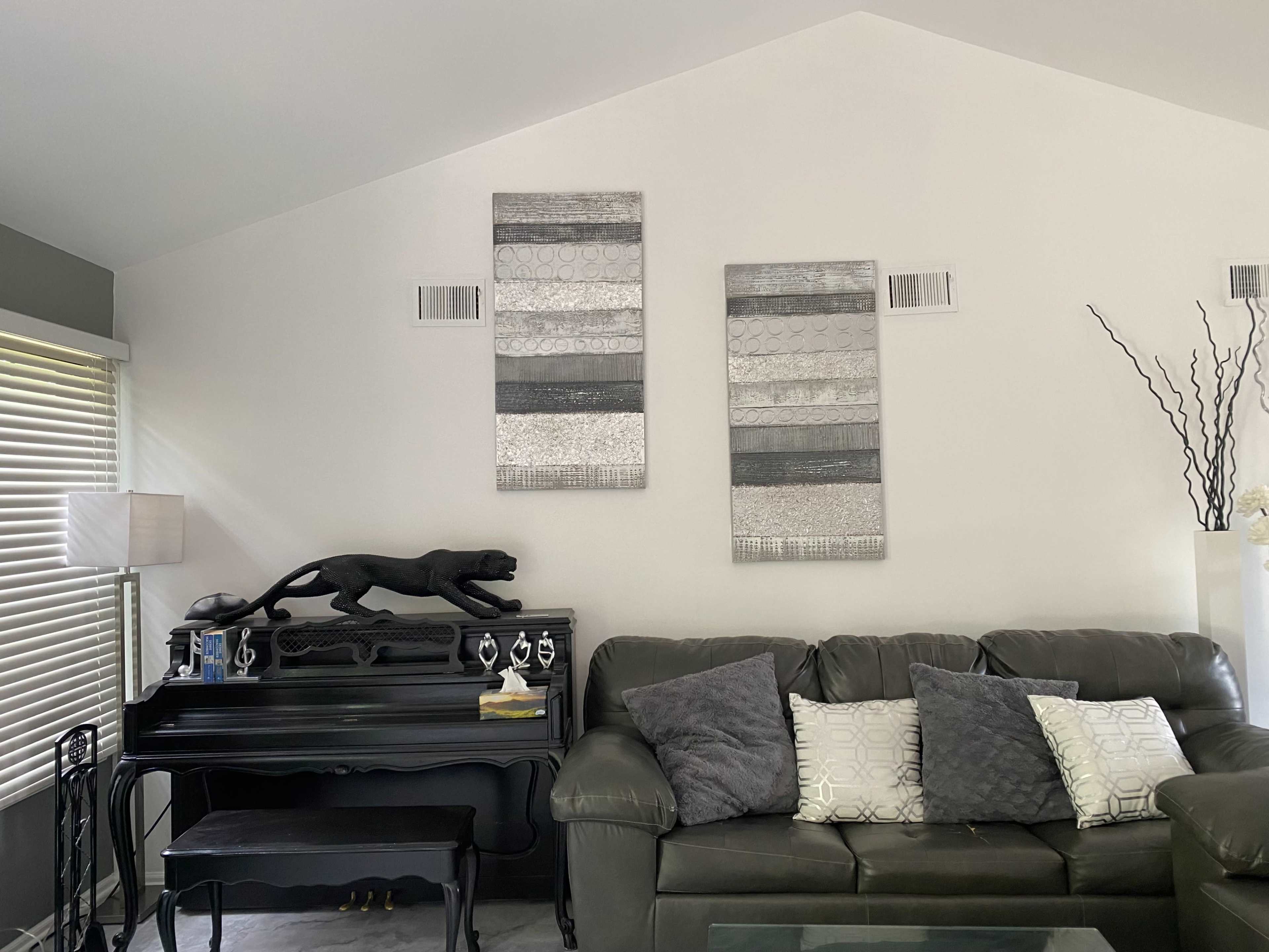 A living room features a black piano with a decorative panther statue on top, accompanied by two silver wall art pieces, and a dark green couch adorned with several pillows.