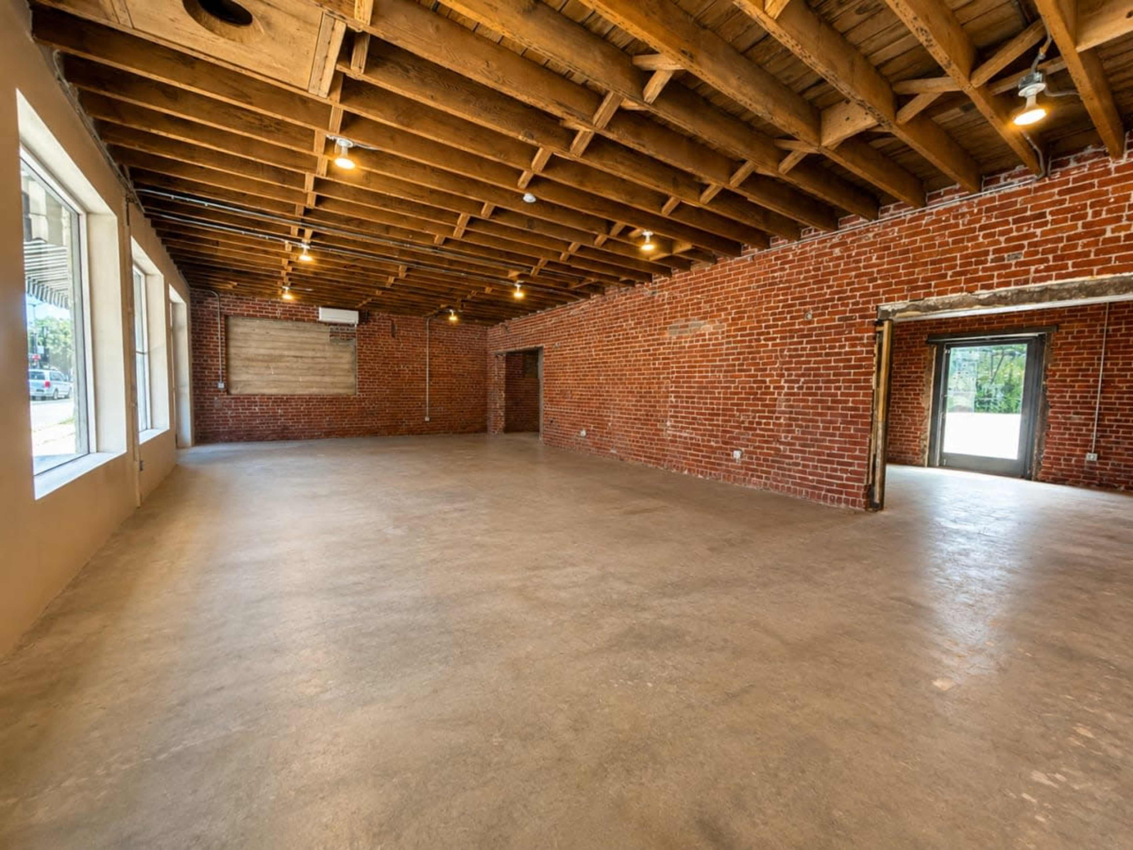 The image shows a spacious indoor area with exposed brick walls, a ceiling with wooden beams, and a concrete floor.
