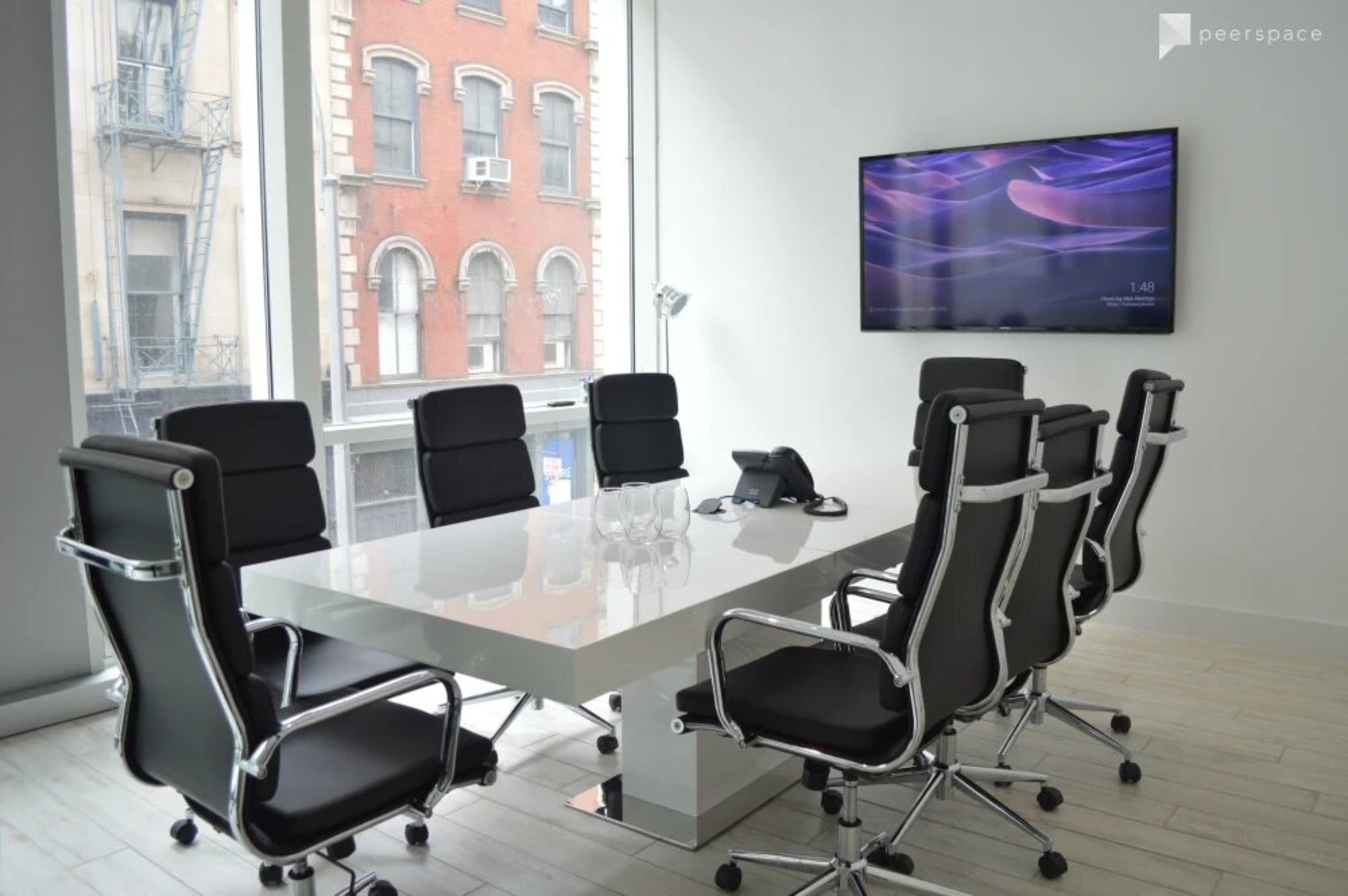 A modern conference room features a large white table surrounded by black ergonomic chairs, with a wall-mounted screen displaying time in the background.