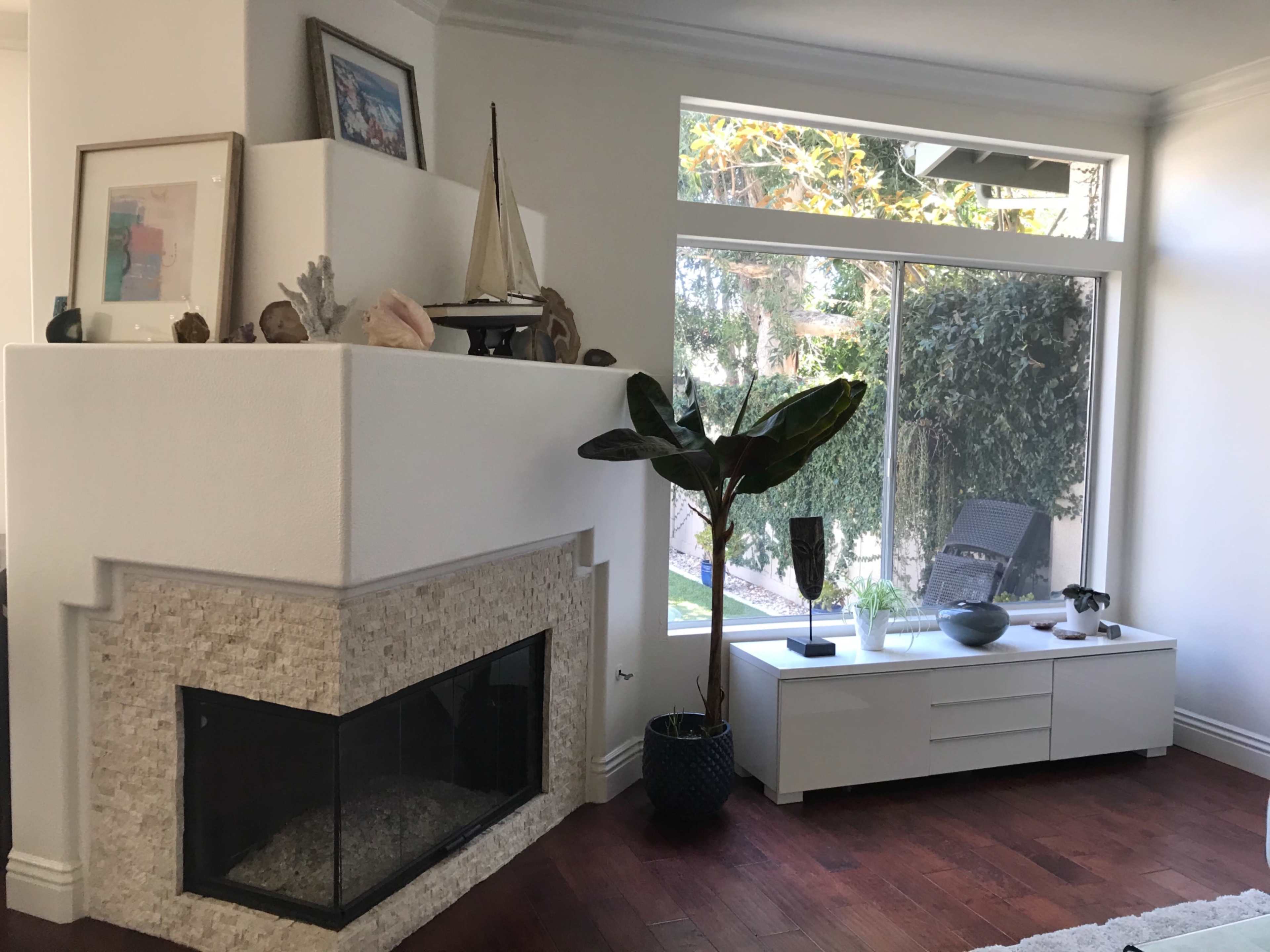 A modern living room with a white fireplace, a large window overlooking greenery, and a minimalist coffee table adorned with decorative items.