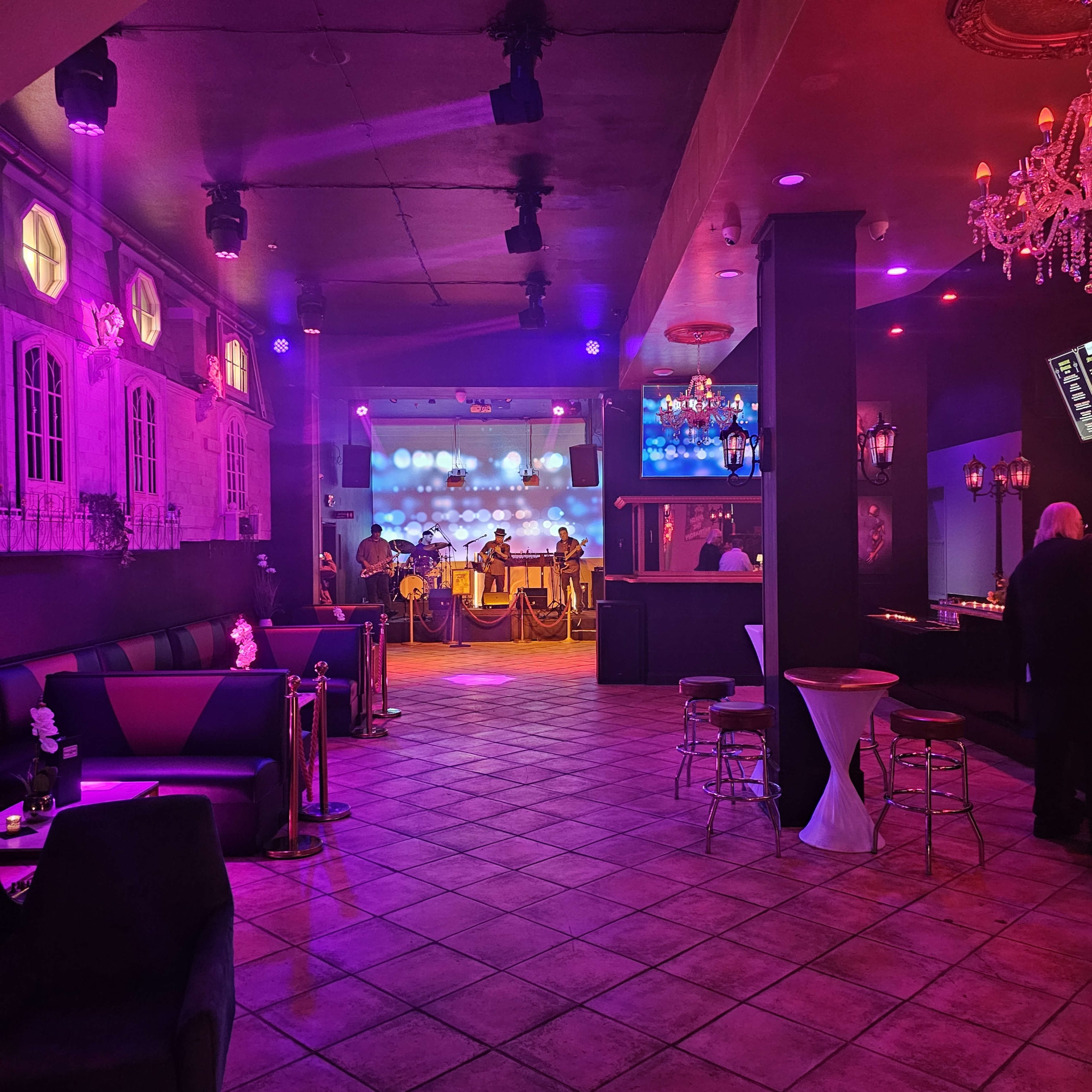 The image shows a dimly lit lounge with a stage for live music, colorful lighting, and seating areas including tables and couches.
