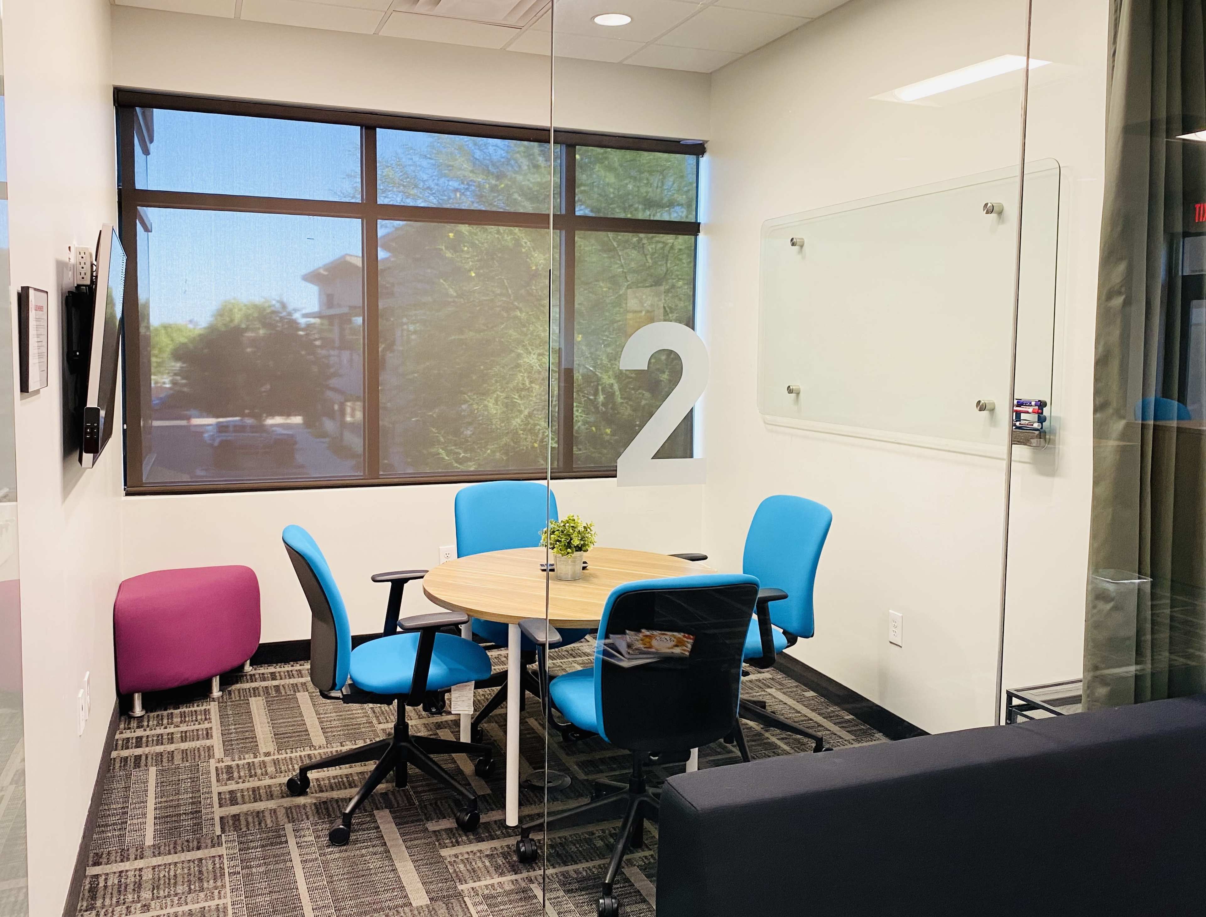 The image shows a small meeting room with a round table and four chairs, a potted plant on the table, and a large window letting in natural light.