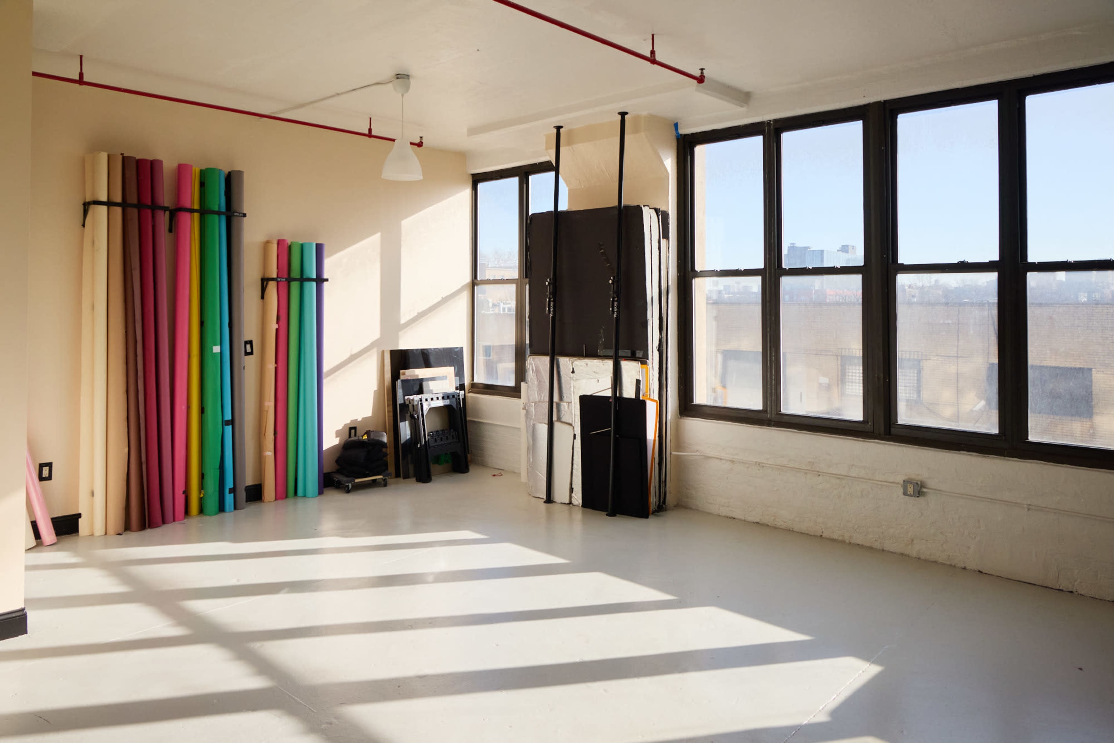 The image shows an empty room with large windows, colorful rolls of paper stacked against one wall, and various equipment leaning against another.
