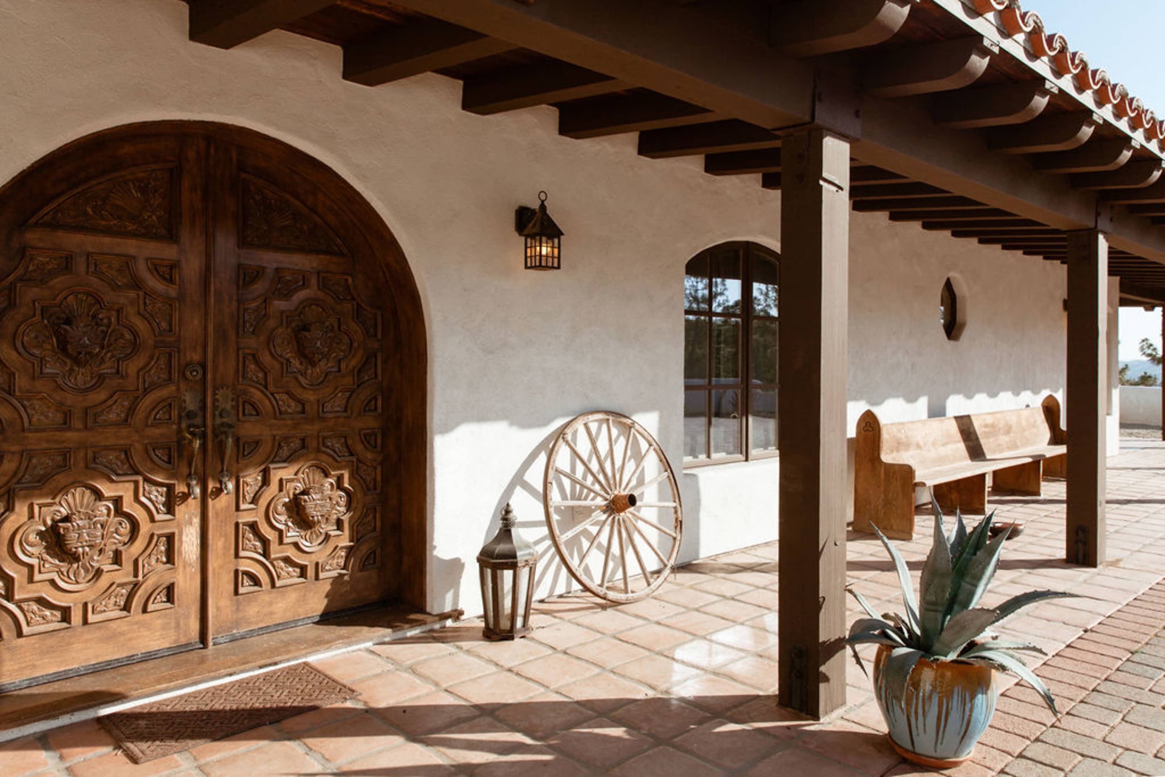 A rustic entrance with a large intricately carved wooden door, a vintage wagon wheel, a potted succulent, and a wooden bench under a tiled roof.