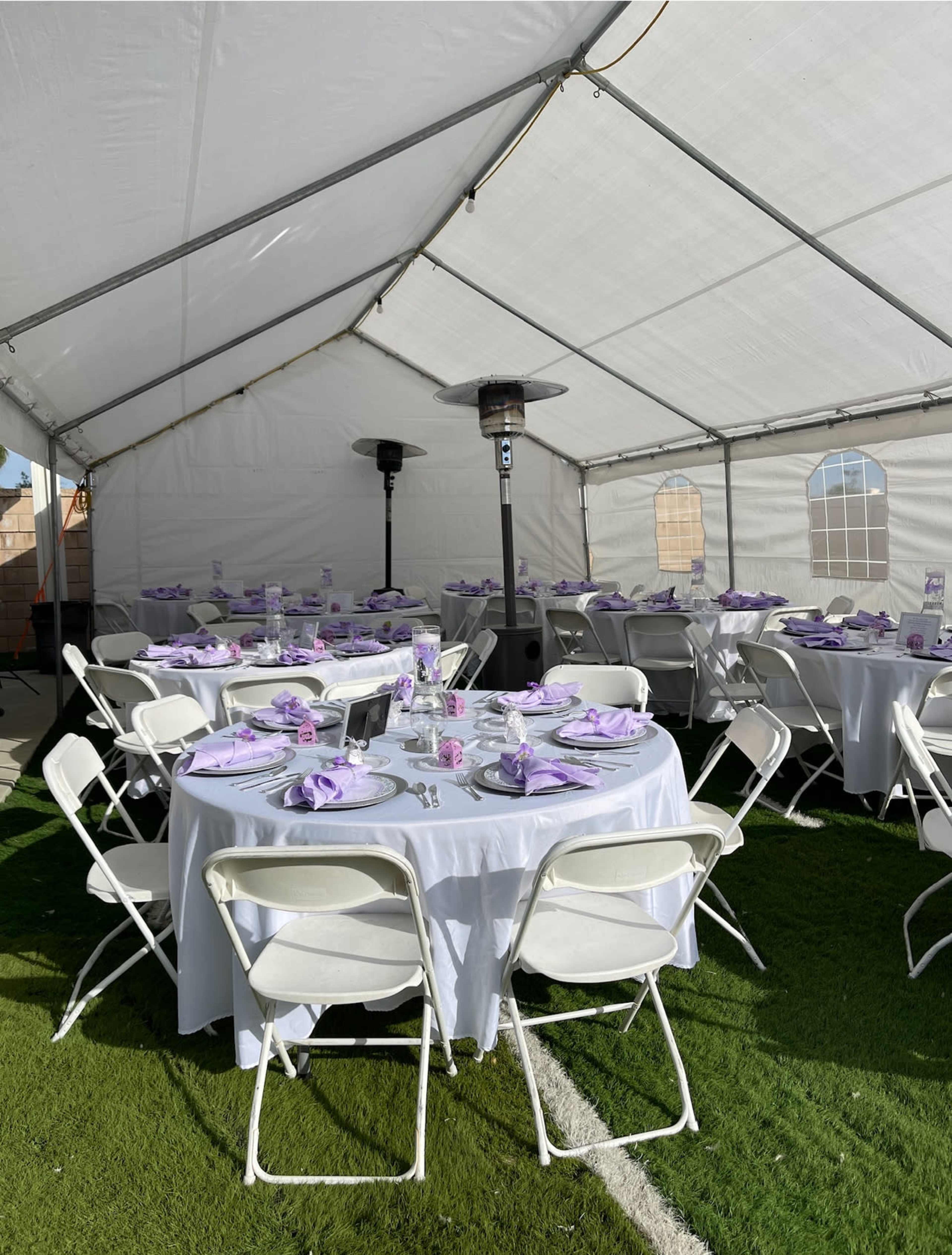 A large tent is set up with several round tables covered in white tablecloths, each adorned with lavender napkins and set for dining.