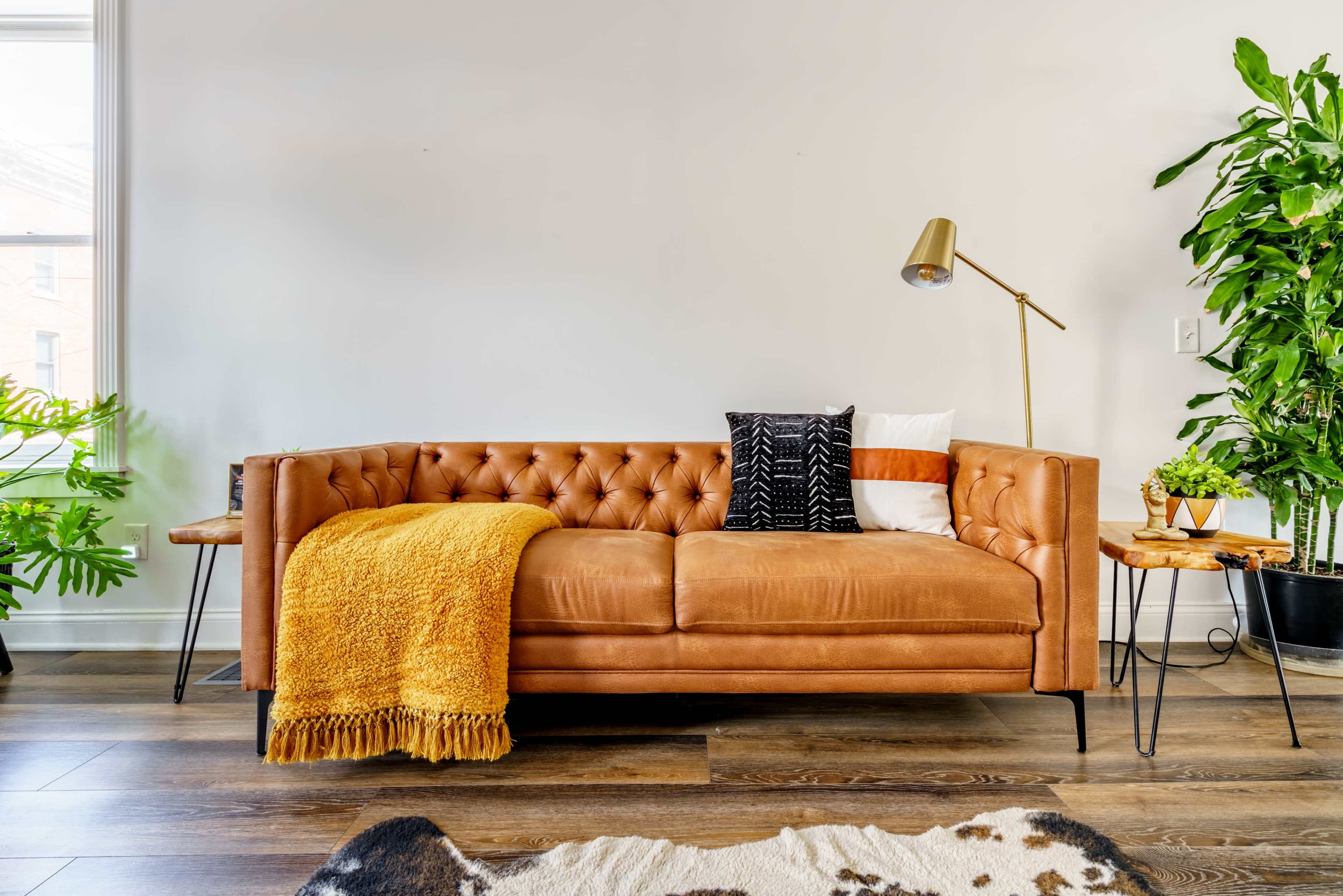 A tufted tan sofa is positioned beside a tall indoor plant and two wooden side tables, accented by decorative pillows and a mustard yellow throw blanket.