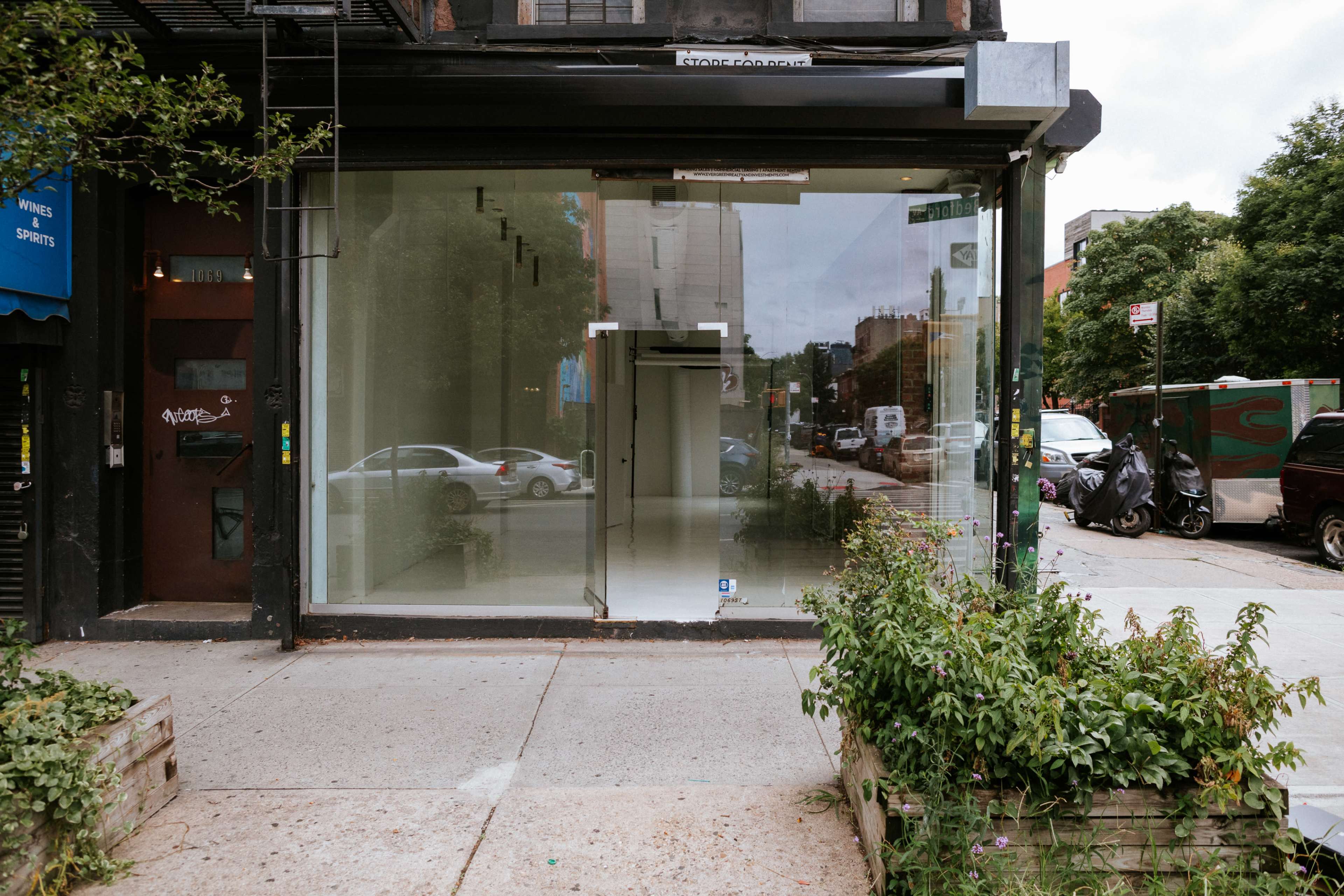 The image shows a vacant storefront with large glass windows facing the street, surrounded by greenery and parked vehicles.