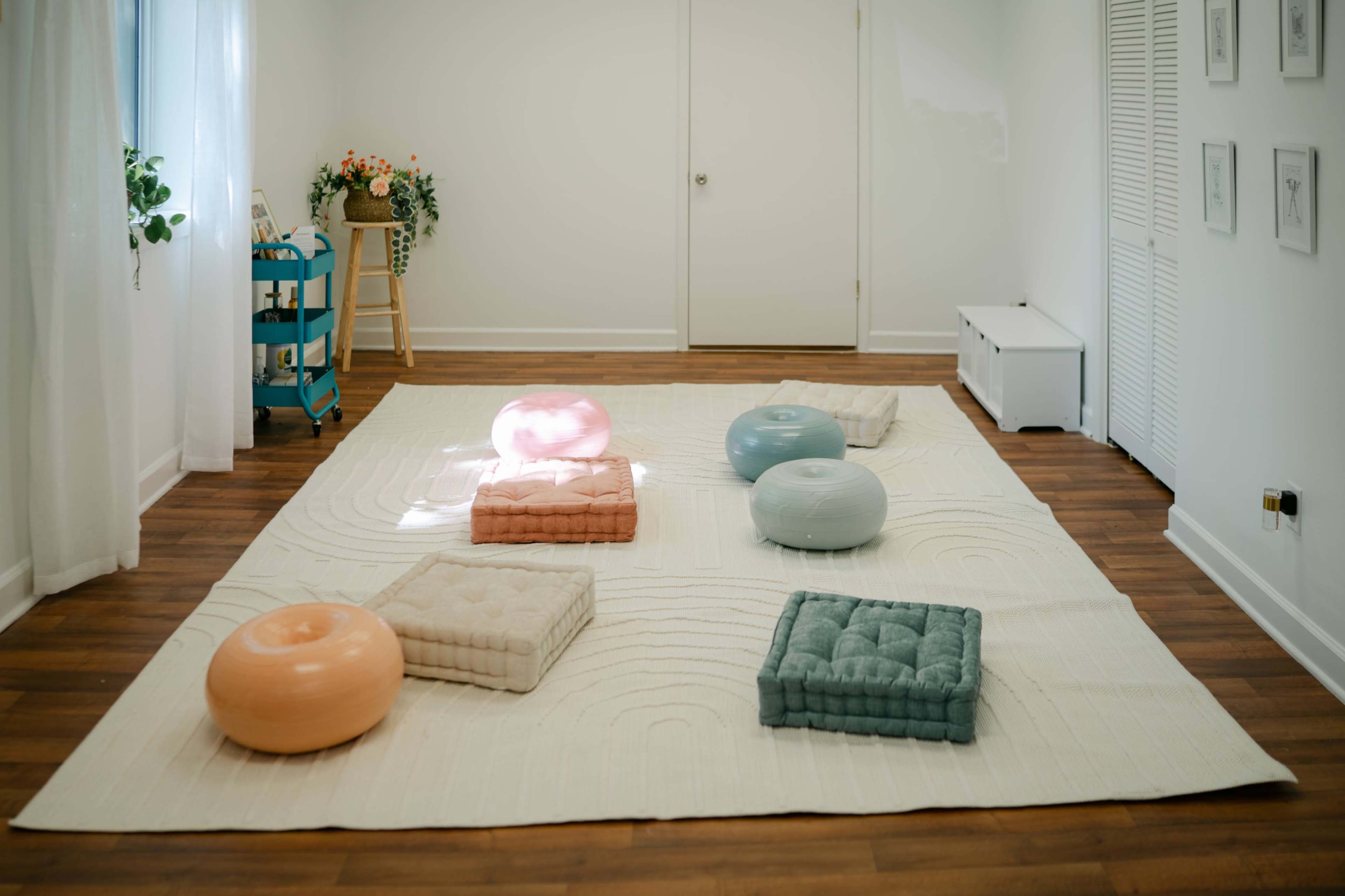 The image shows a spacious room with a light-colored floor, featuring a large rug on which are various round and square cushions in pastel colors.