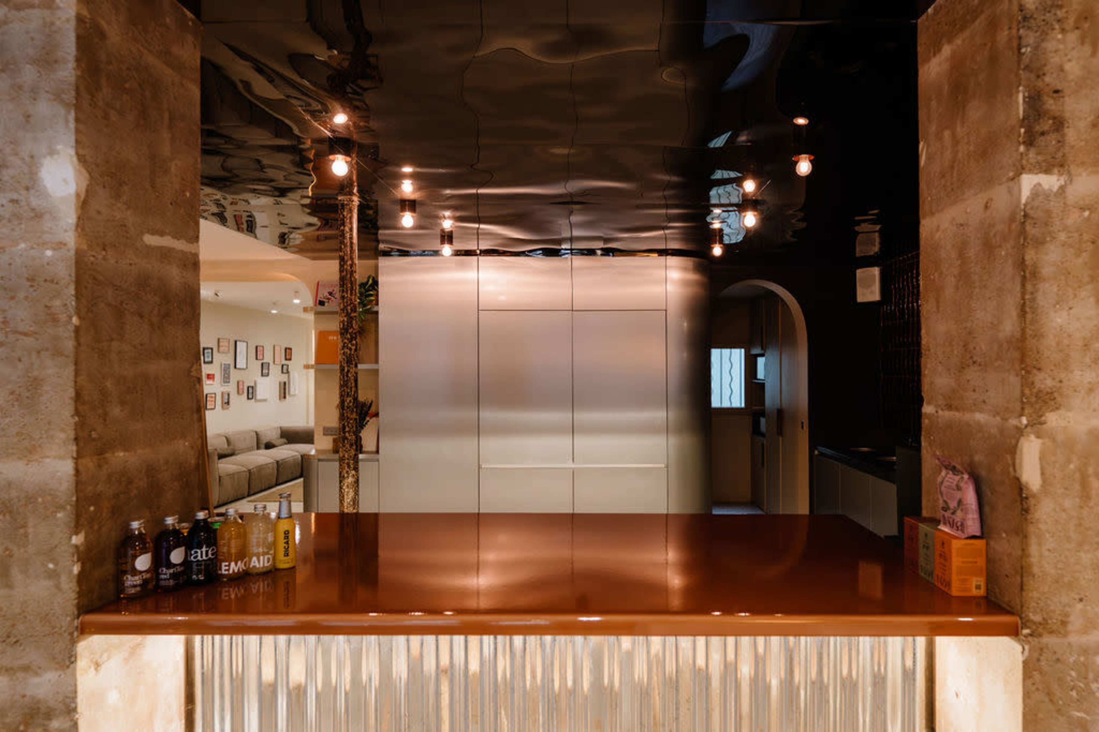 The image shows a modern bar area with a polished wooden counter, a reflective ceiling, and dim hanging lights, set against a backdrop of concrete walls.