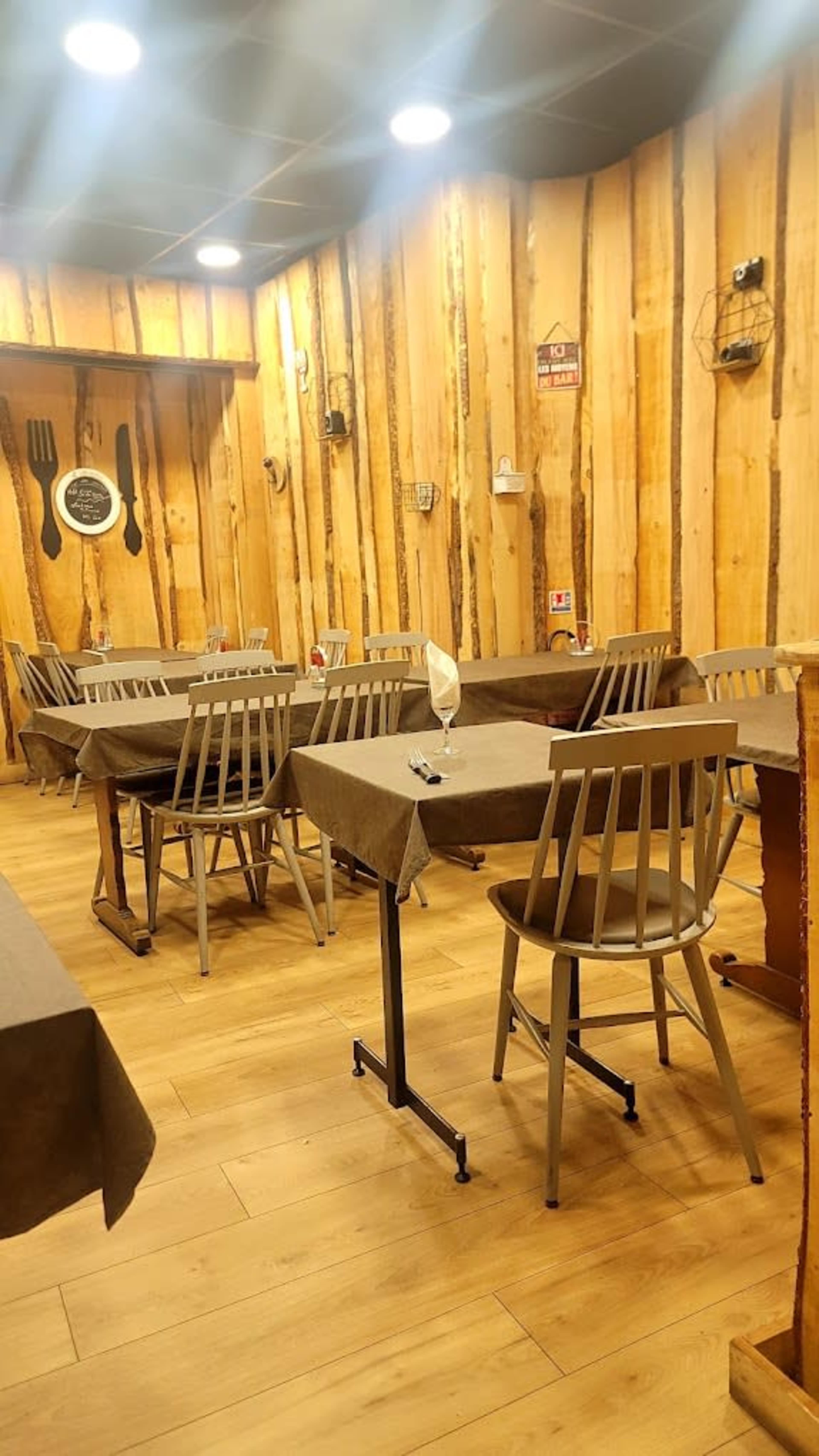 The image depicts a restaurant interior featuring wooden walls, neatly arranged tables with brown tablecloths, and simple wooden chairs.