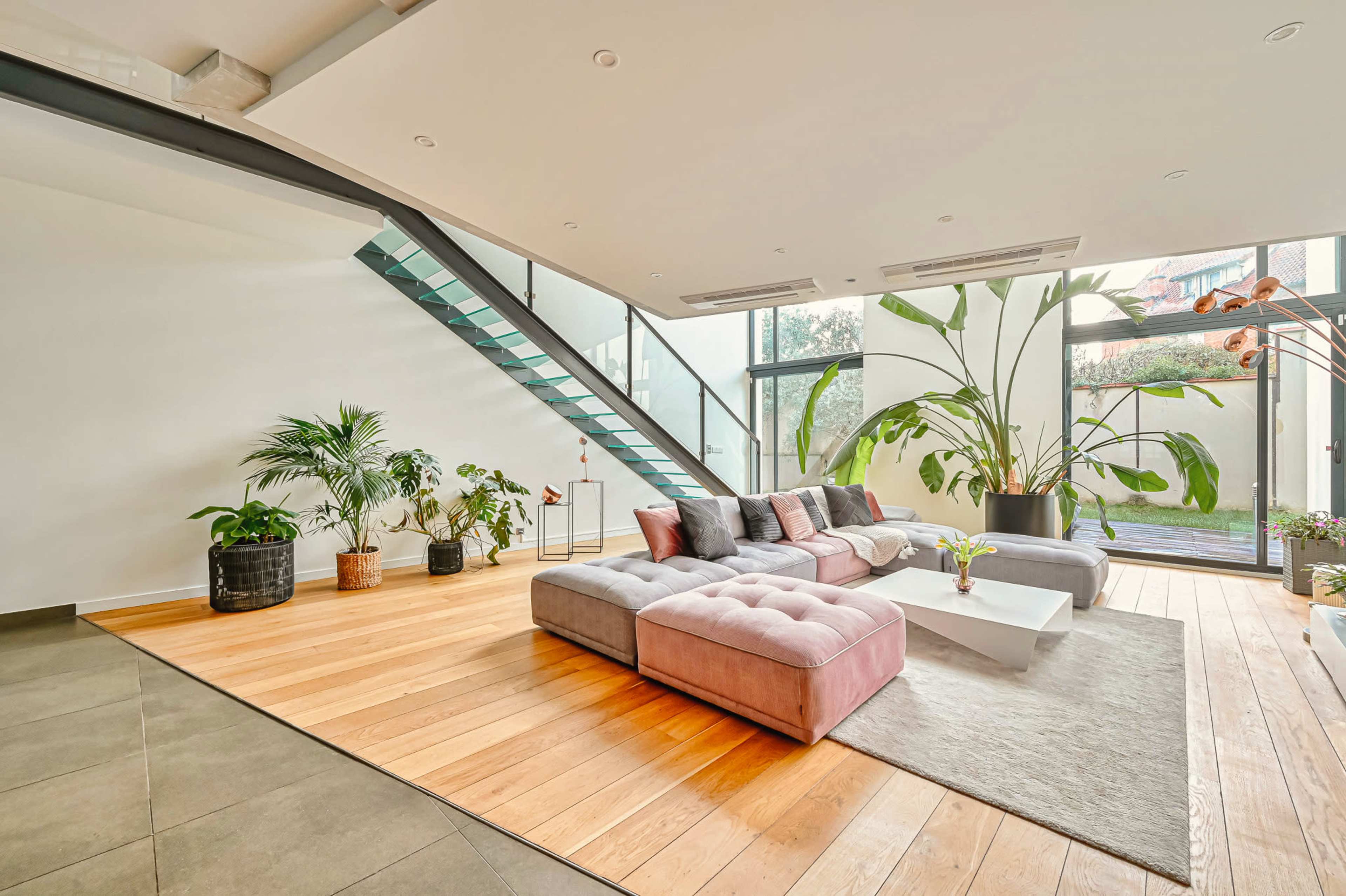 A spacious living room features a modern sectional sofa, large windows, and a staircase with a glass railing.
