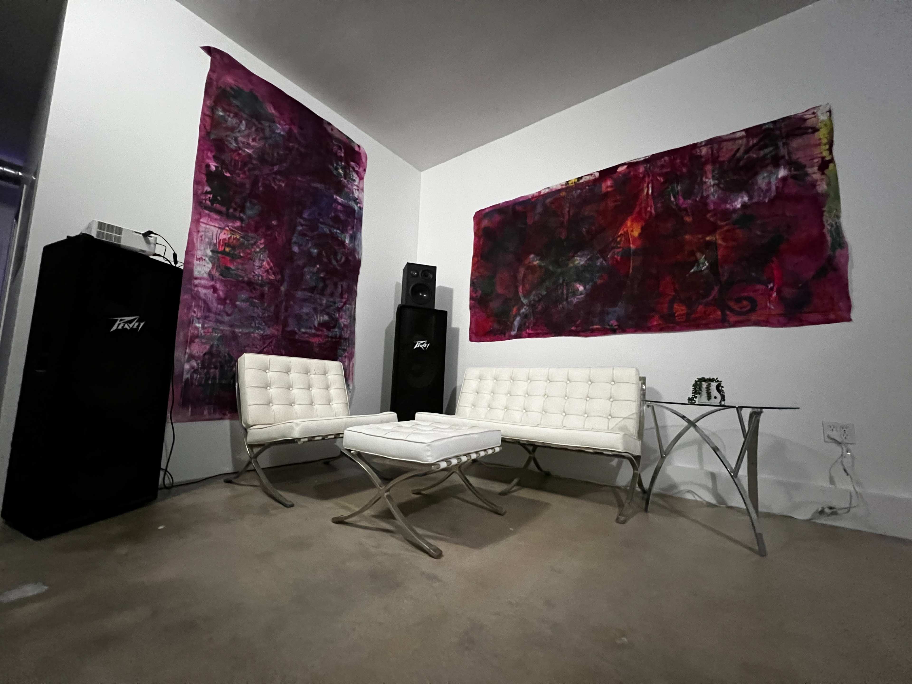 A minimalist seating area with two white chairs and a coffee table against a background of large, colorful abstract paintings and a black speaker.