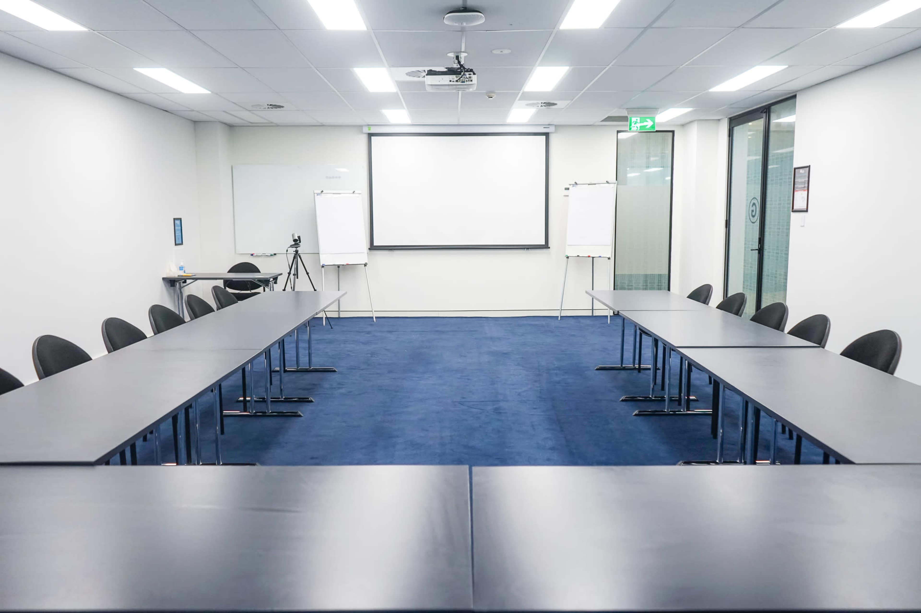 A modern conference room features a large rectangular table surrounded by chairs, with a projector screen and whiteboards visible at one end.