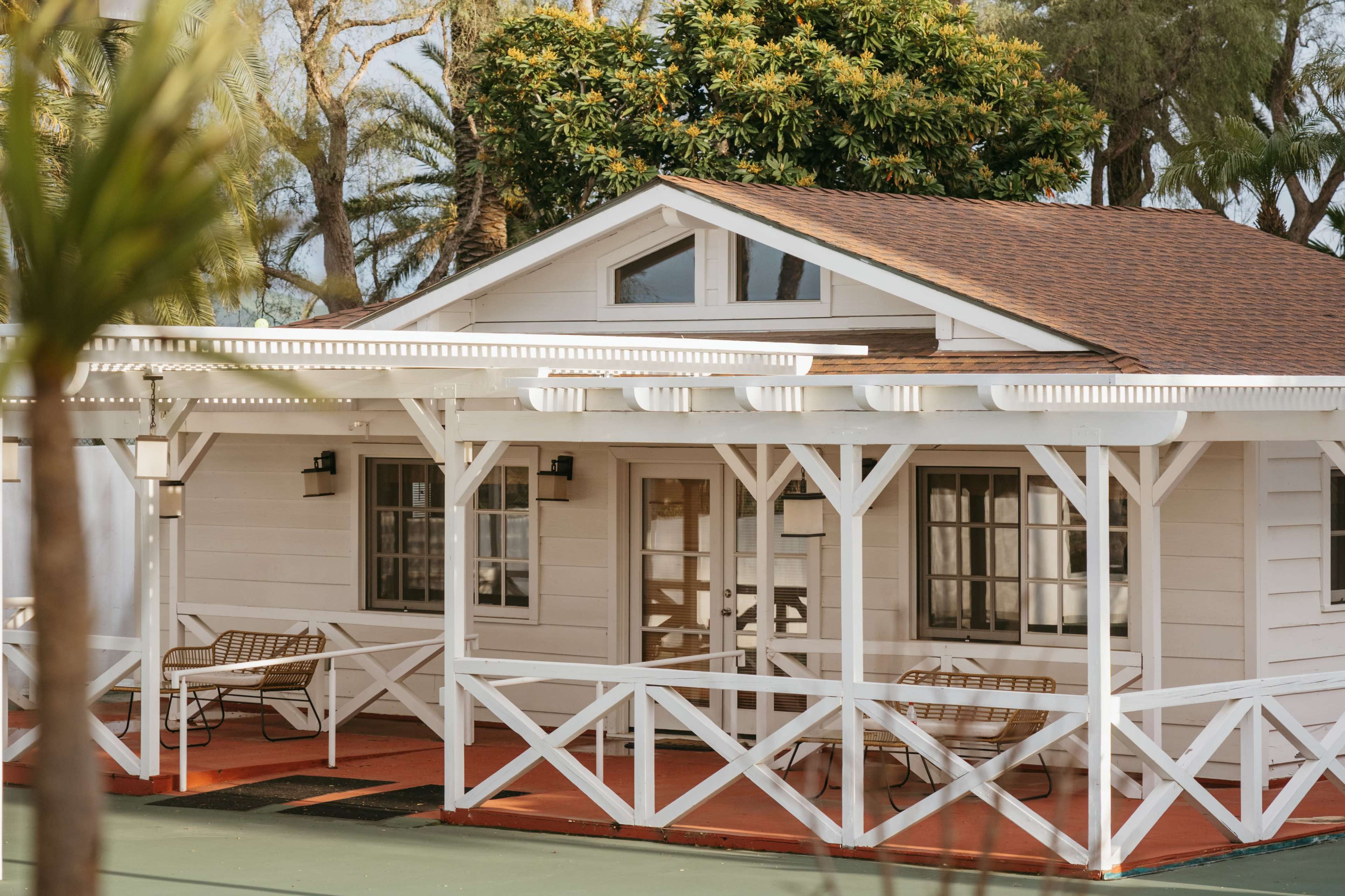 A small white cottage with a brown roof features a wraparound porch surrounded by greenery.
