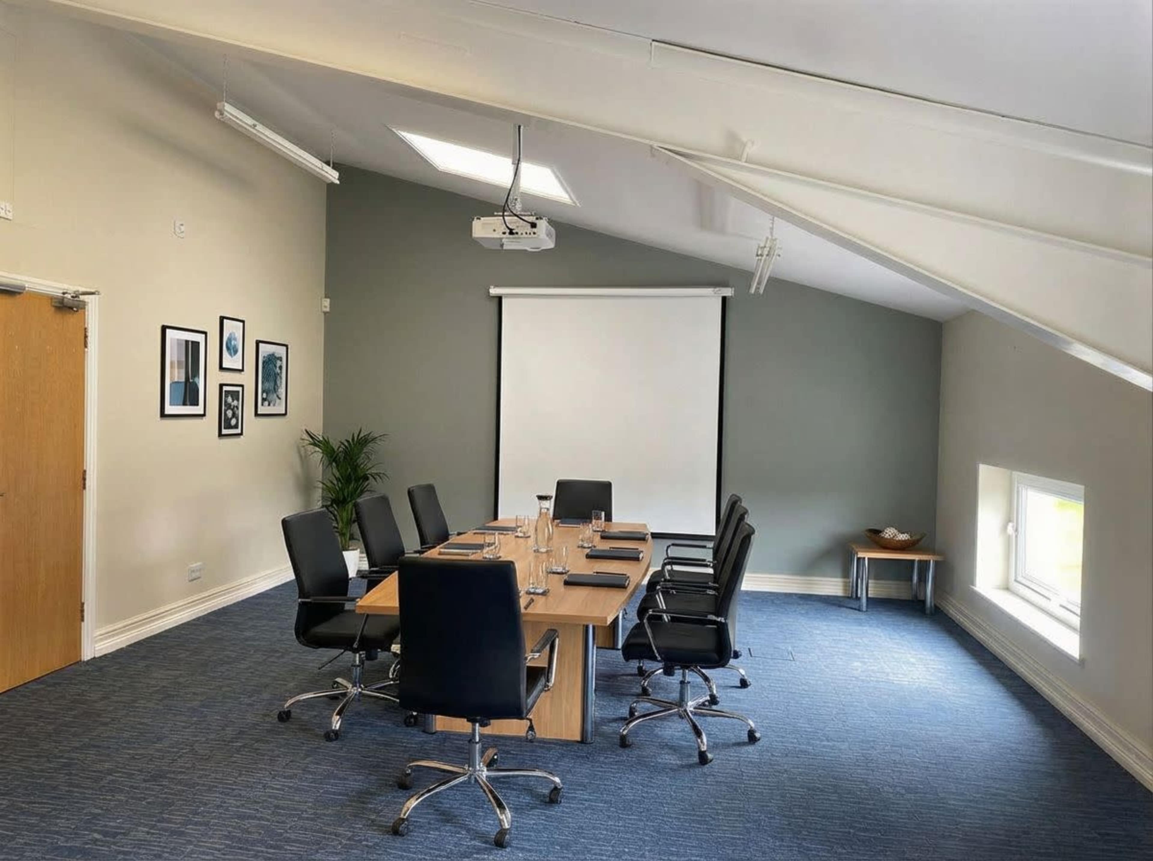The image shows a modern meeting room with a long wooden table, black chairs, a projector, and a large screen on the wall.