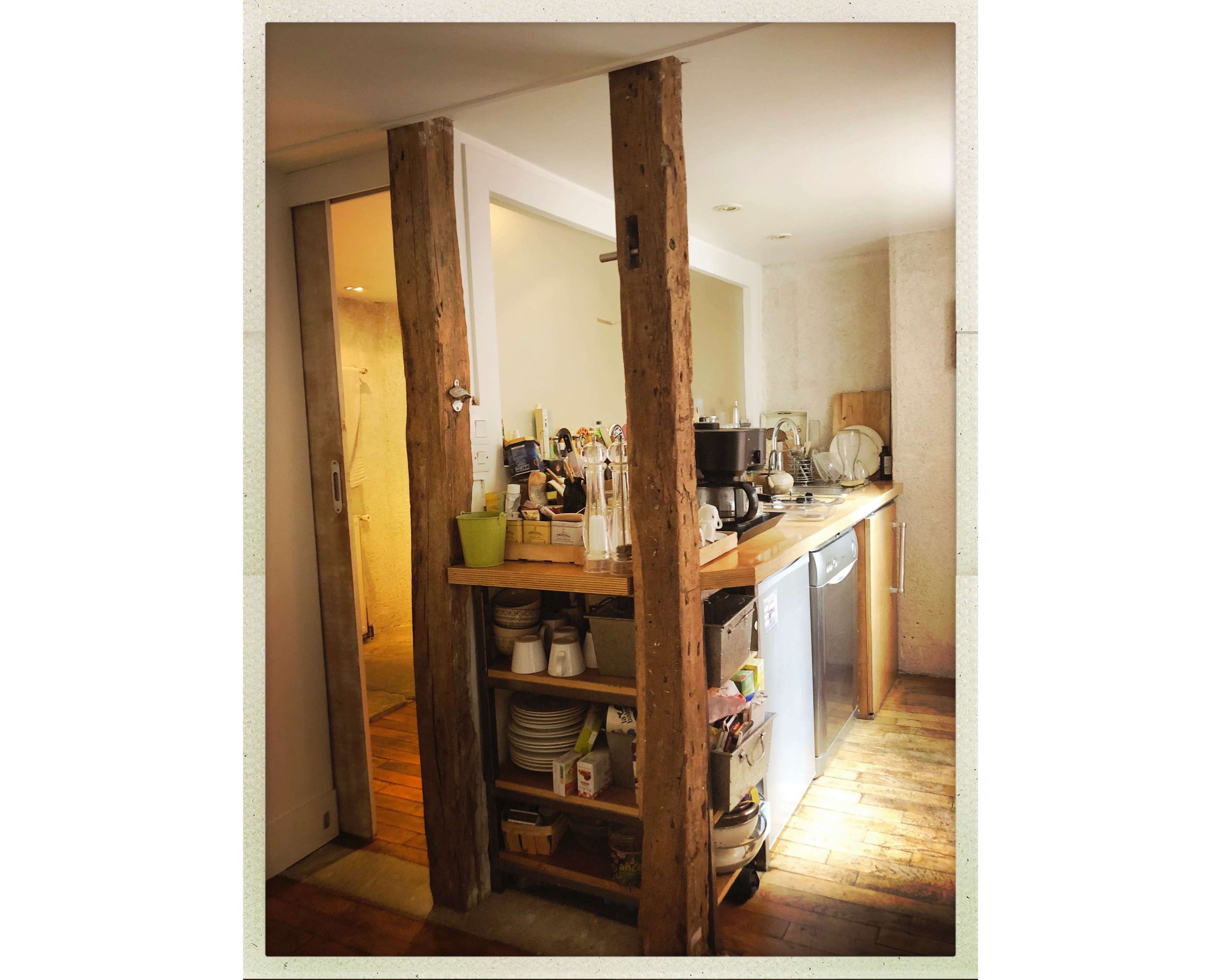 The image shows a kitchen with wooden beams on either side, featuring a countertop filled with kitchen appliances, utensils, and dishes, along with an open doorway leading to a bathroom area.