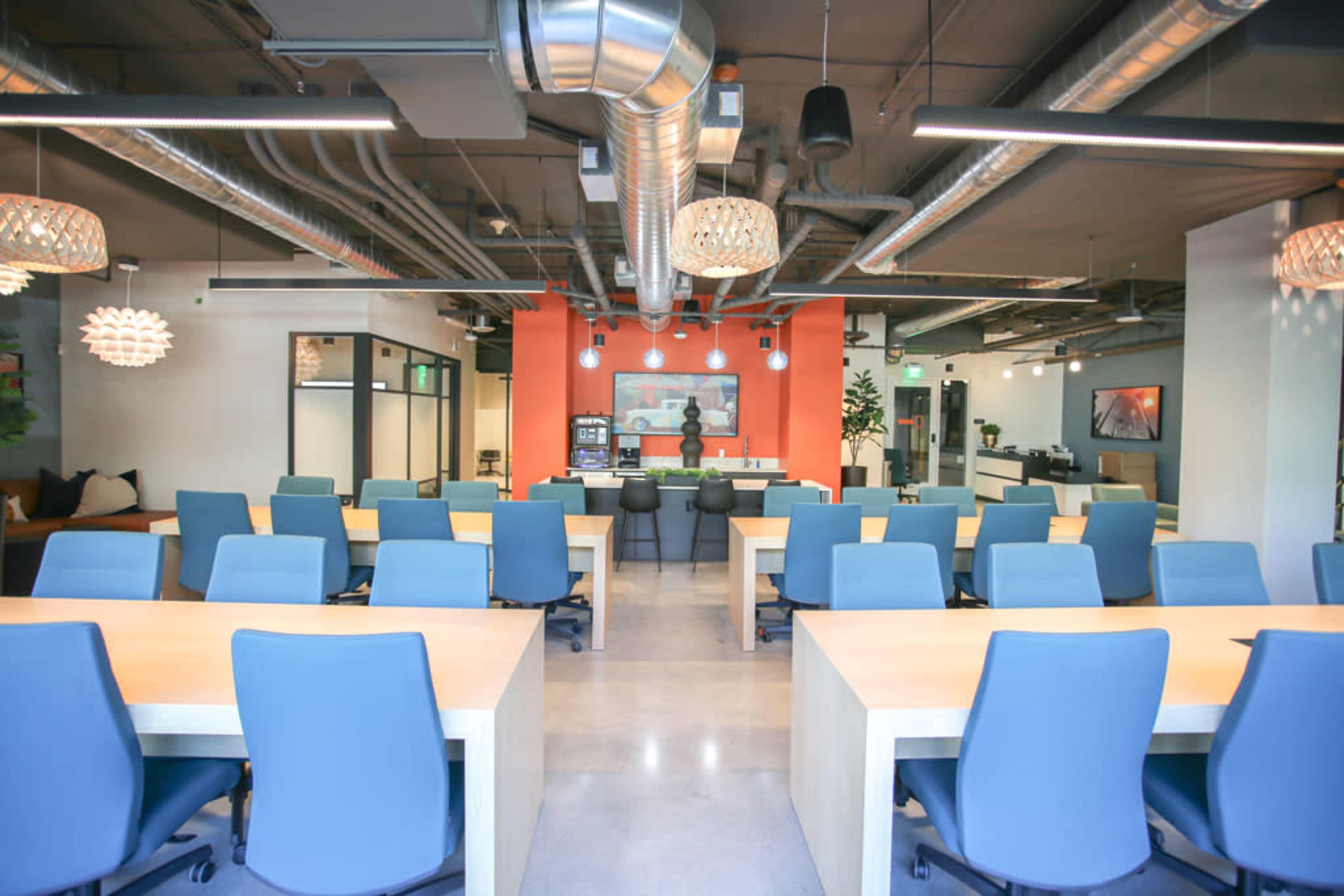 A modern office space with several rows of blue chairs and light wooden desks, and features a vibrant orange accent wall.