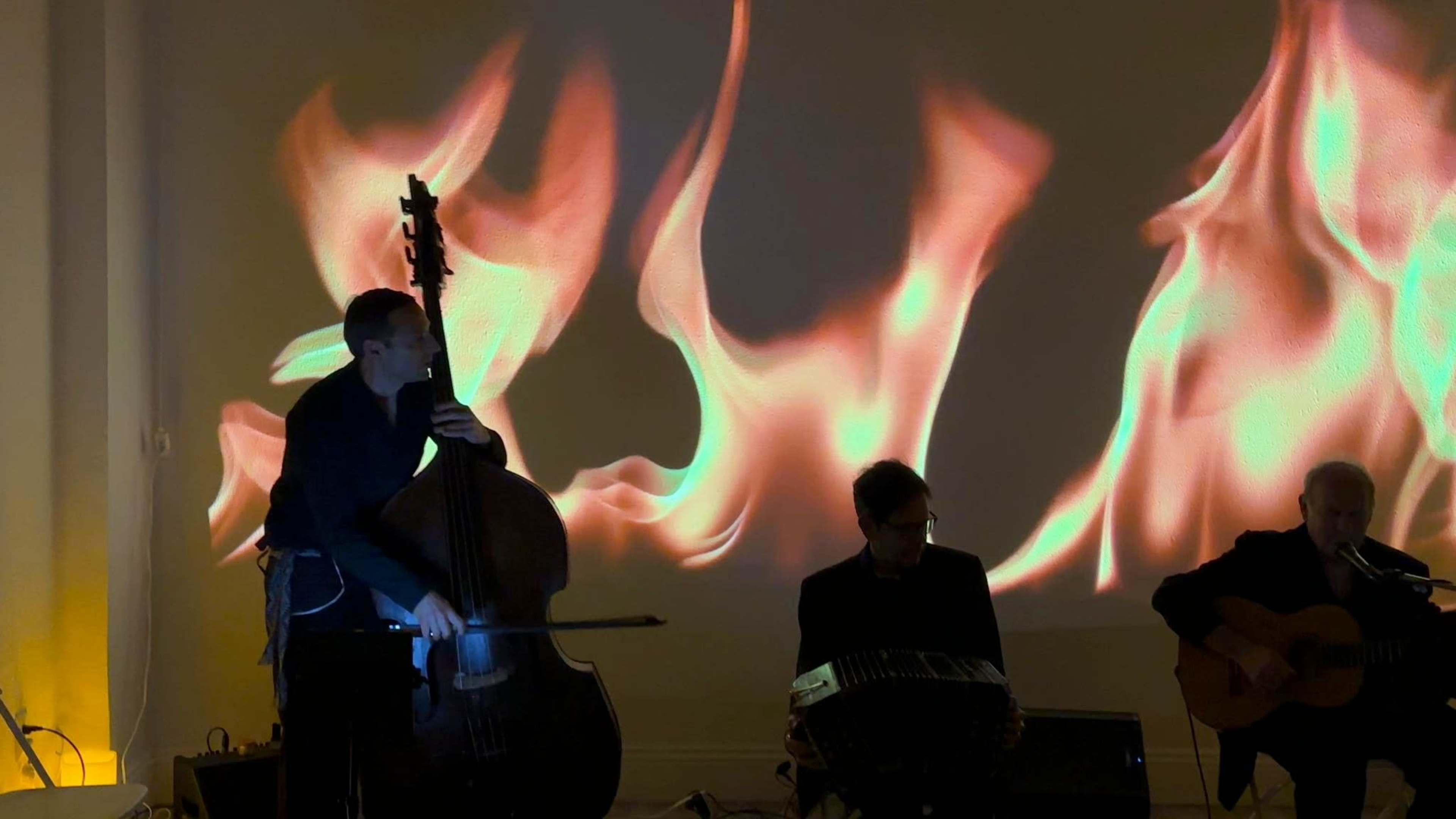 A musician plays a double bass in front of a backdrop featuring animated flames, while two other musicians accompany him on guitar and a different instrument.