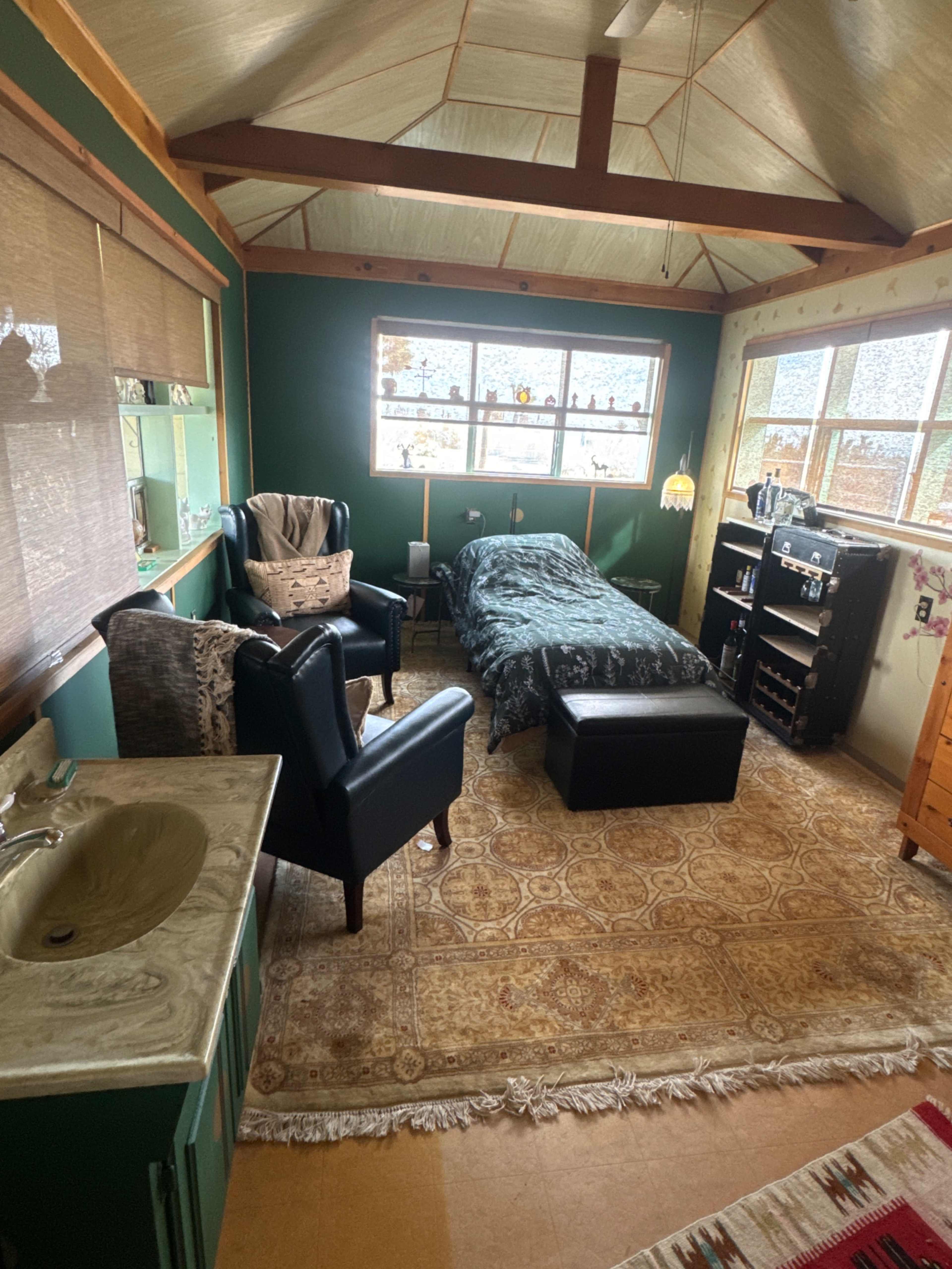 The image shows a small, cozy bedroom with a bed, two chairs, a dresser, and a sink, all featuring a mix of wood and green decor.