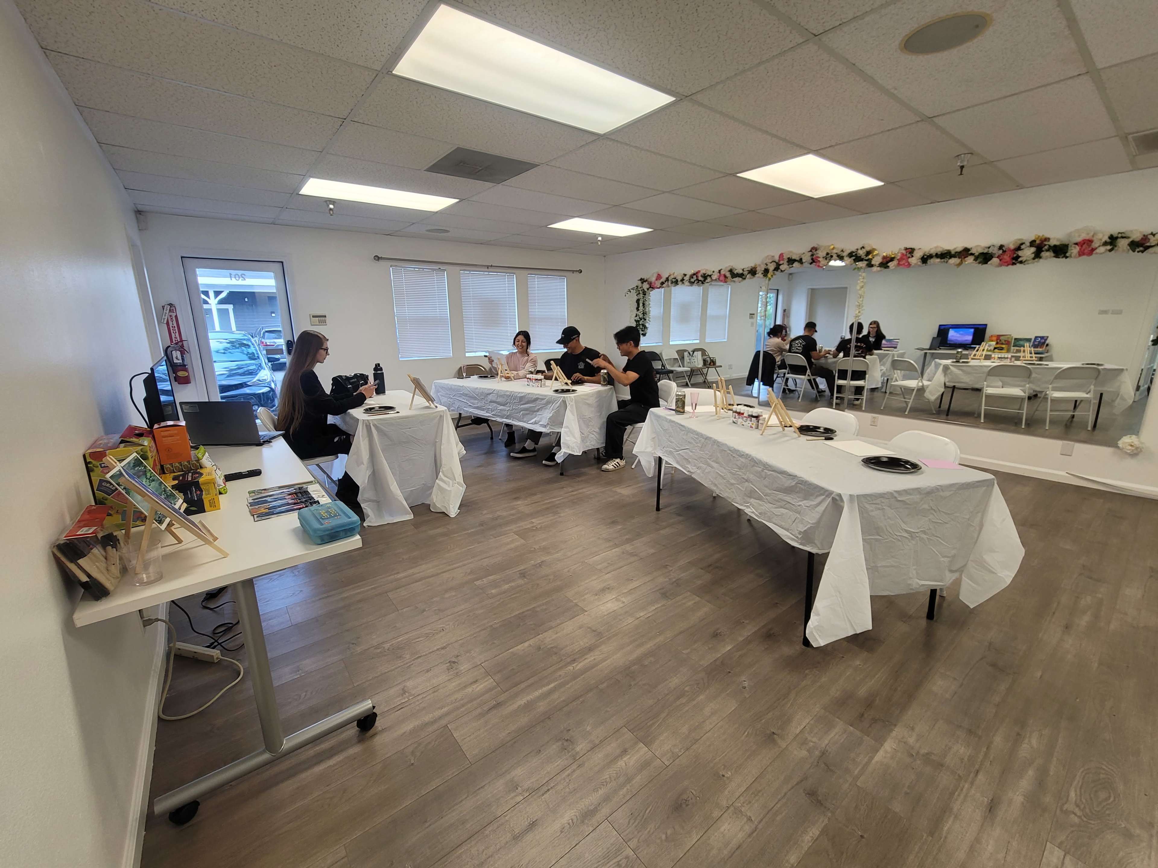 The image shows a spacious, well-lit room with several tables covered in white cloth, where people are engaged in activities, surrounded by decorative elements along the walls.