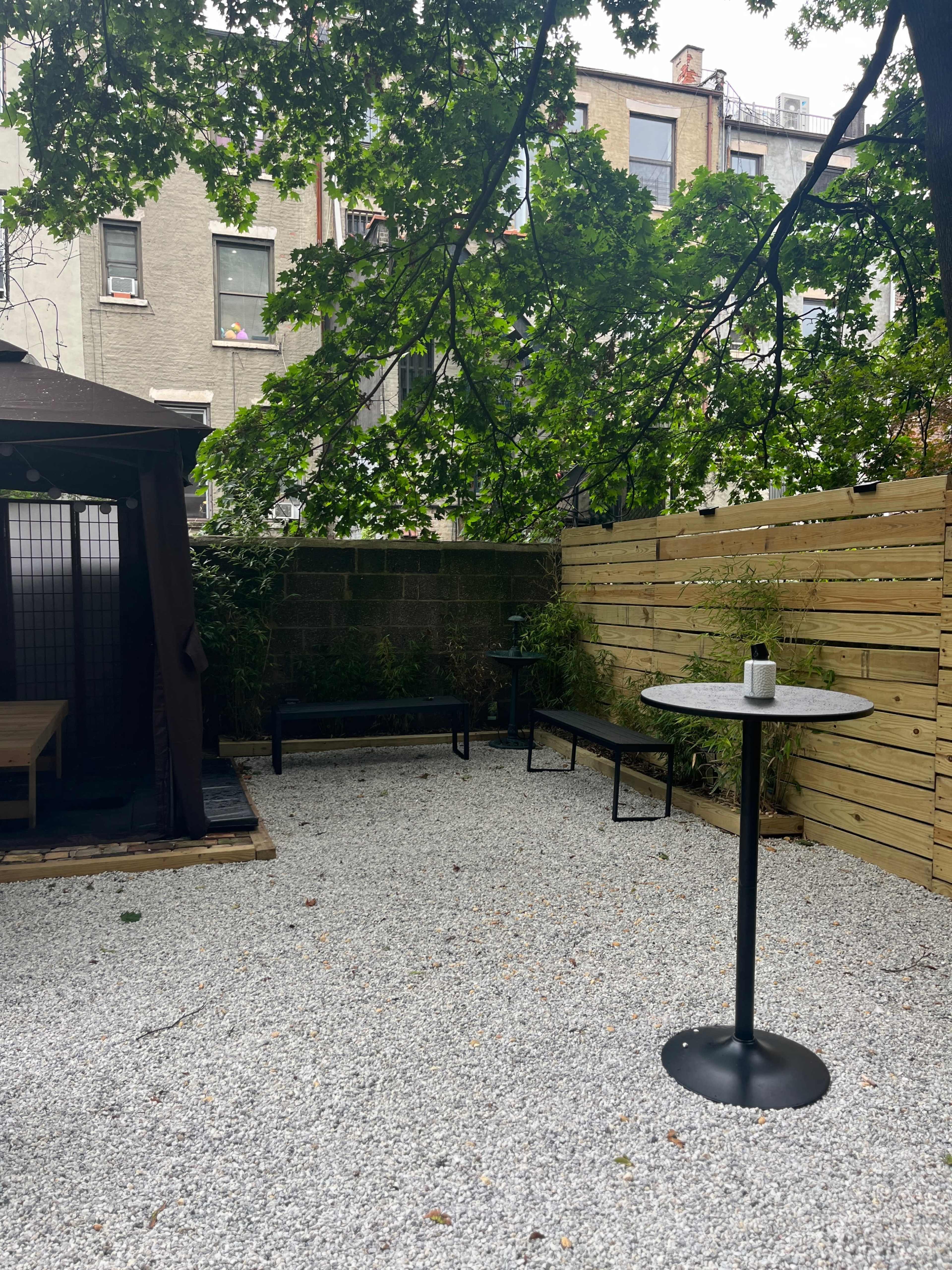 The scene features a gravel courtyard surrounded by wooden fencing and greenery, with a table and chairs under a large tree.