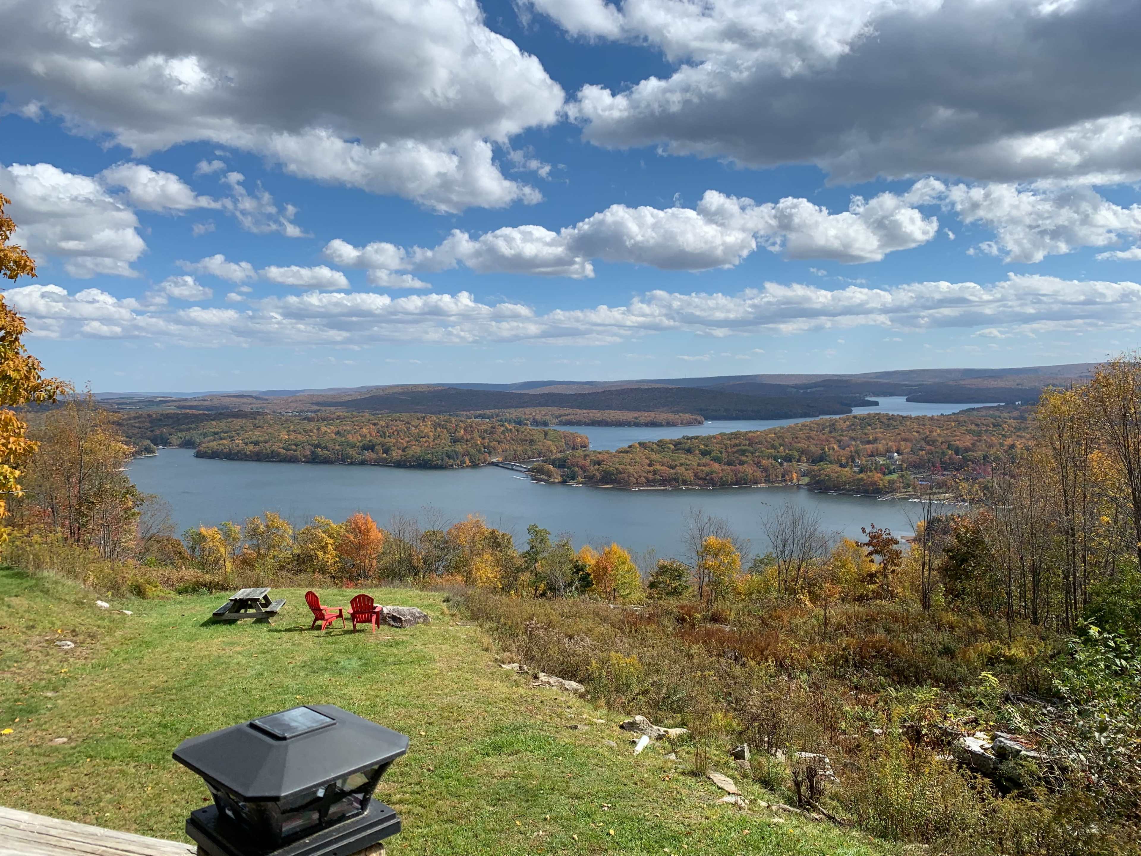 The image shows a scenic view of a lake surrounded by autumn-colored trees from a vantage point, with two red chairs in the foreground.
