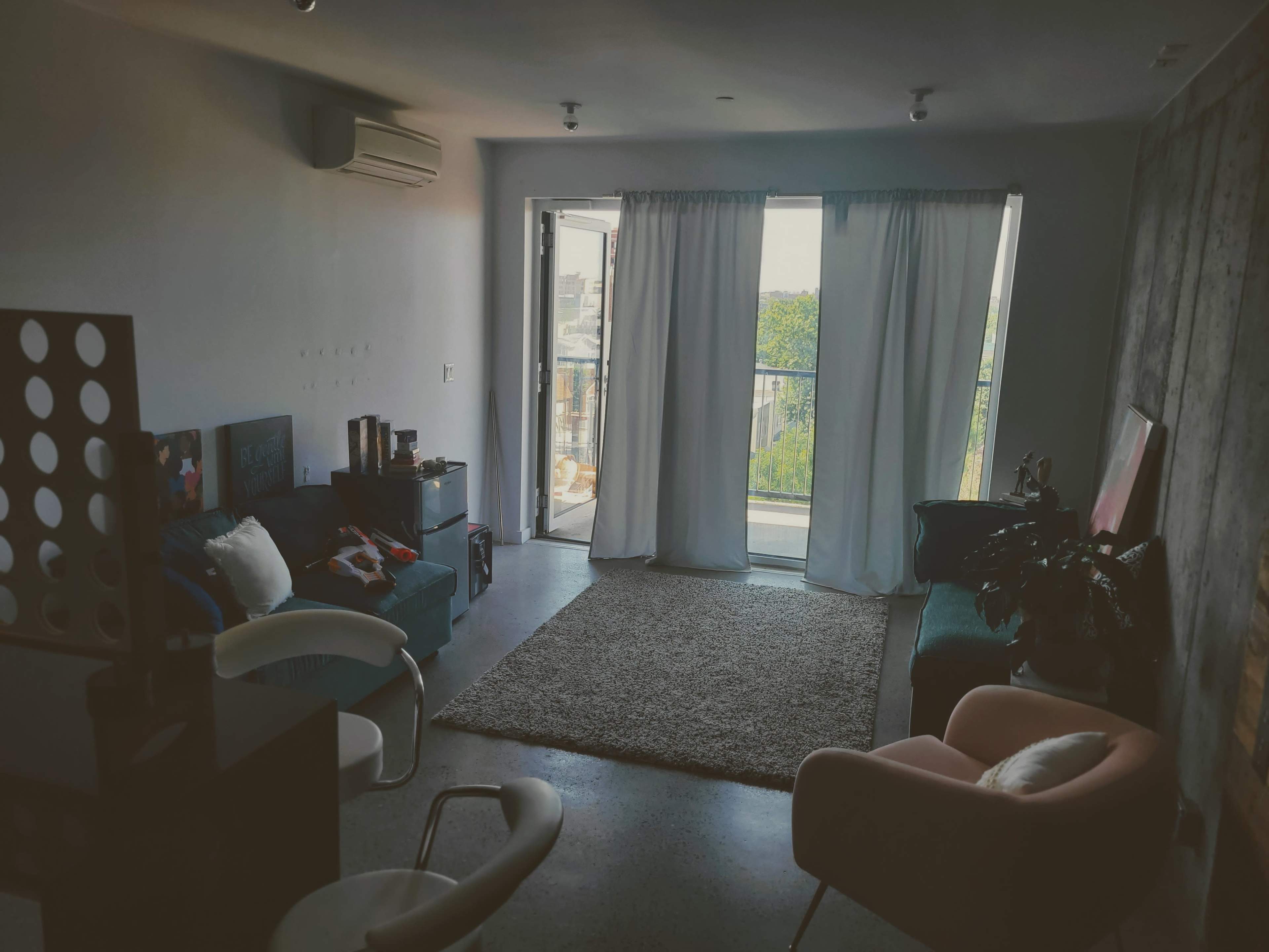 A modern living room with a balcony, featuring a sofa, a chair, and a soft rug, illuminated by natural light.