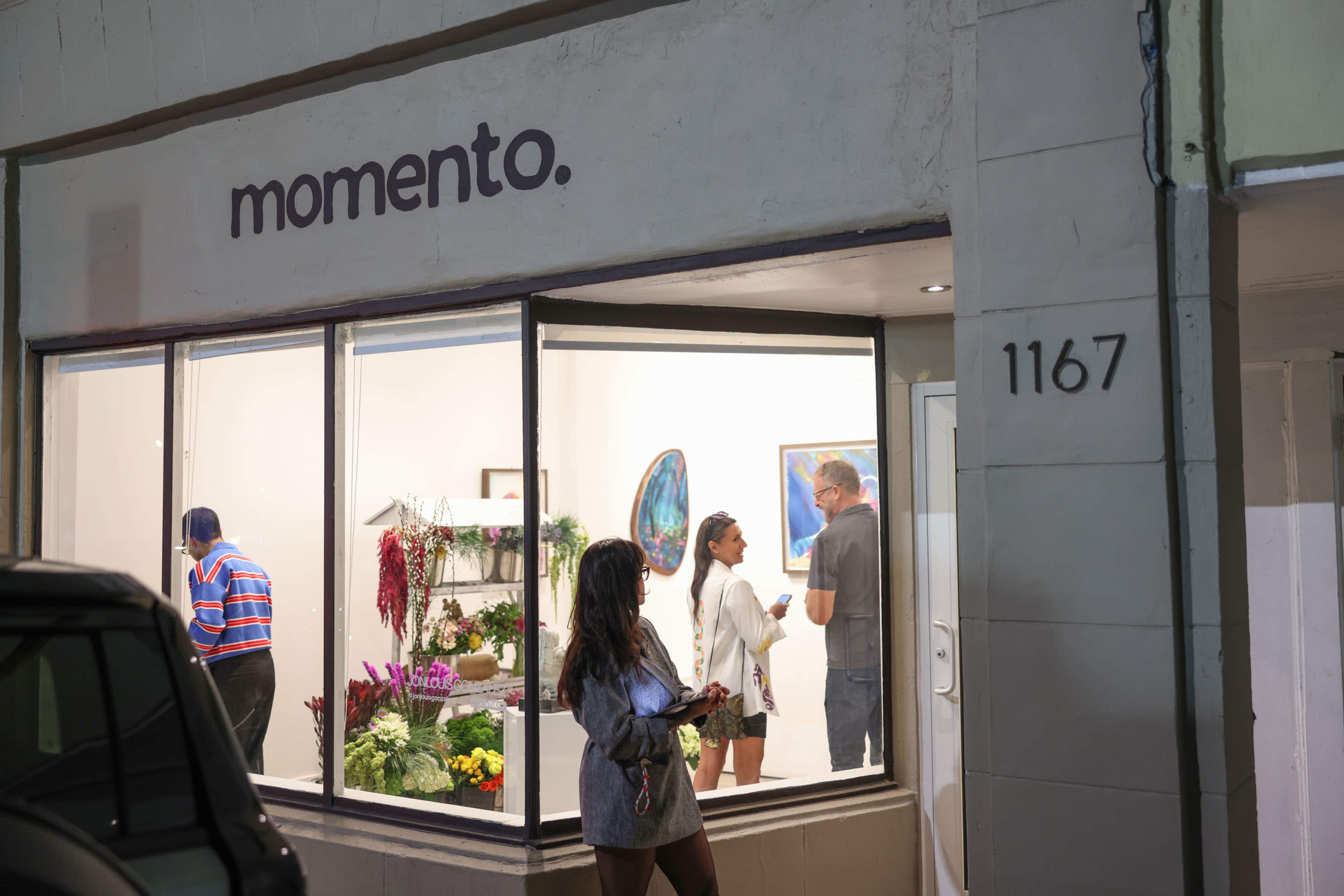 The image shows a gallery named "momento." with people interacting inside and displaying floral arrangements and artworks in the window.