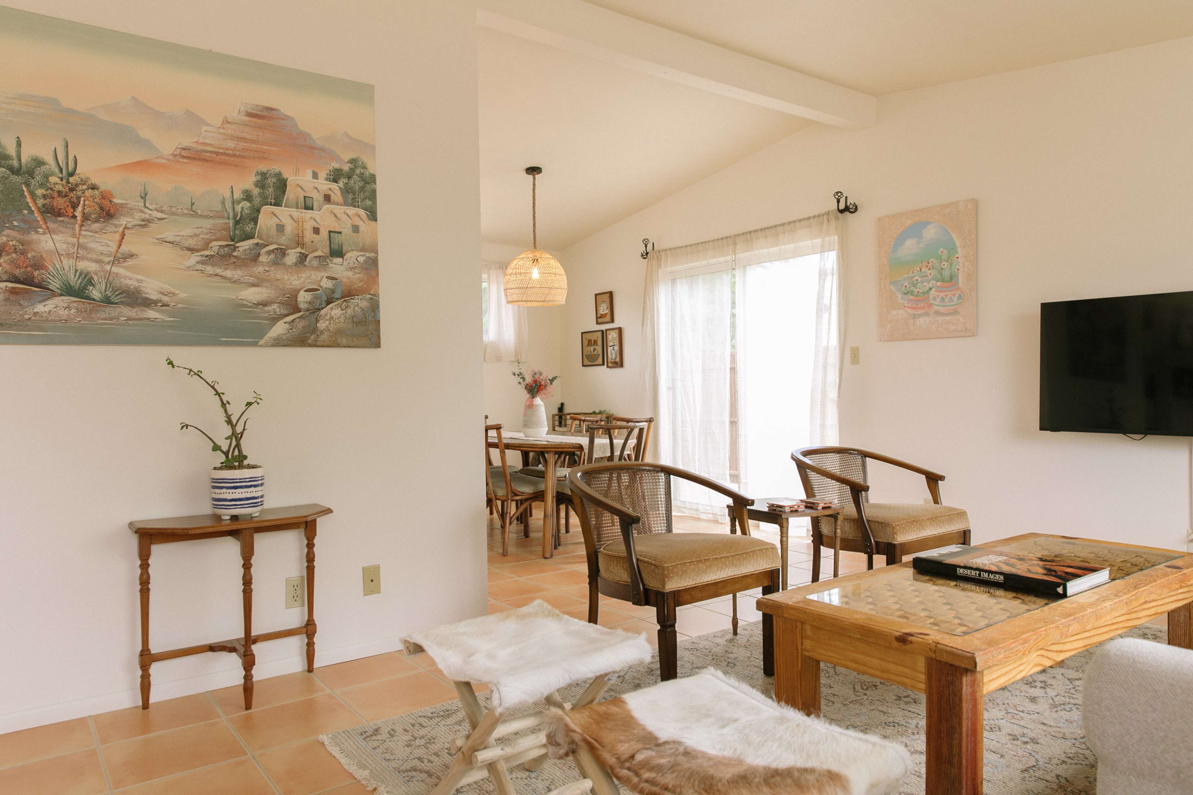 A bright, open living space featuring a wooden coffee table, two chairs, and a wall painting of a desert landscape.
