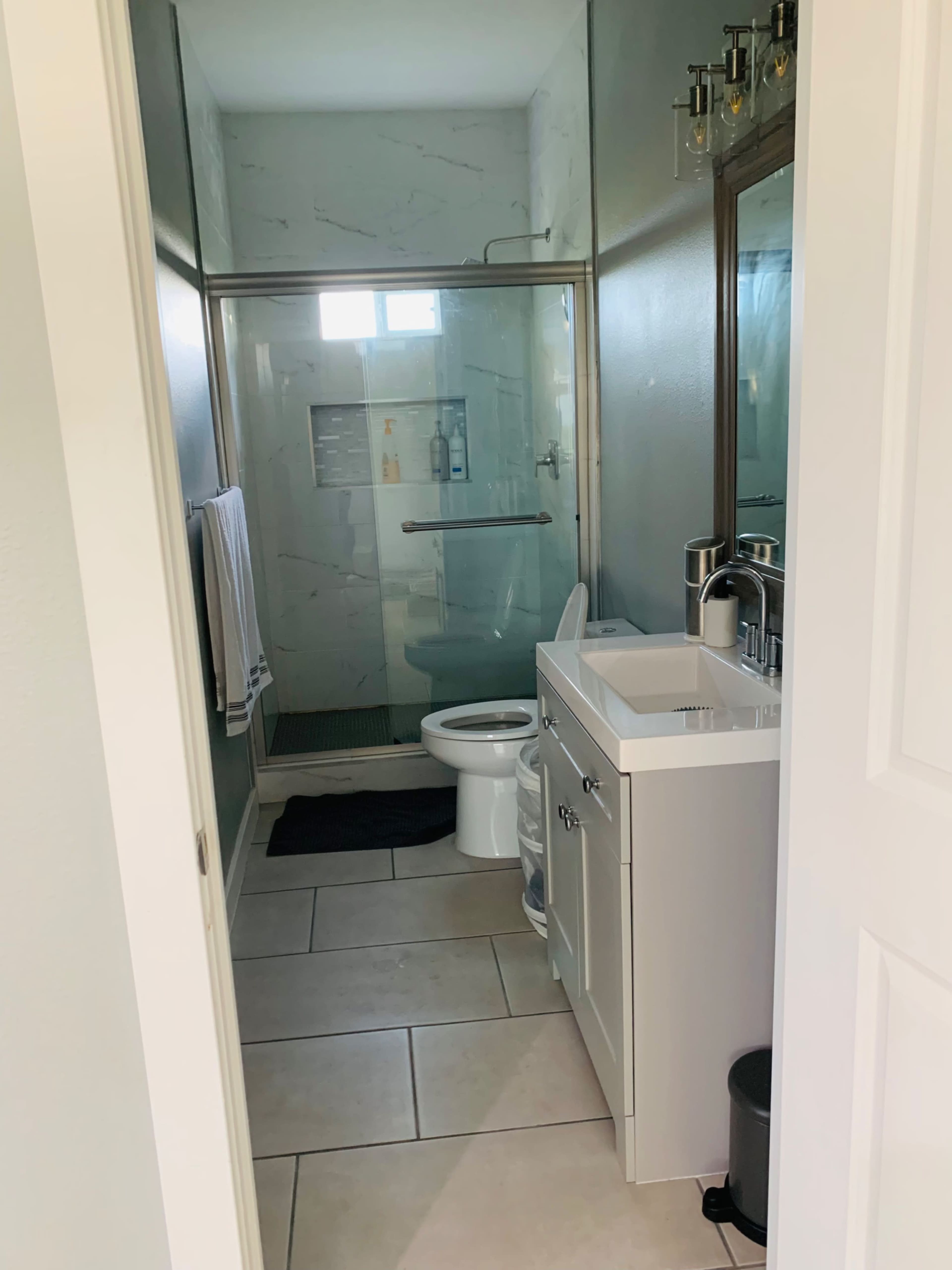 The image shows a modern bathroom with a glass shower, white vanity, toilet, and tile flooring.