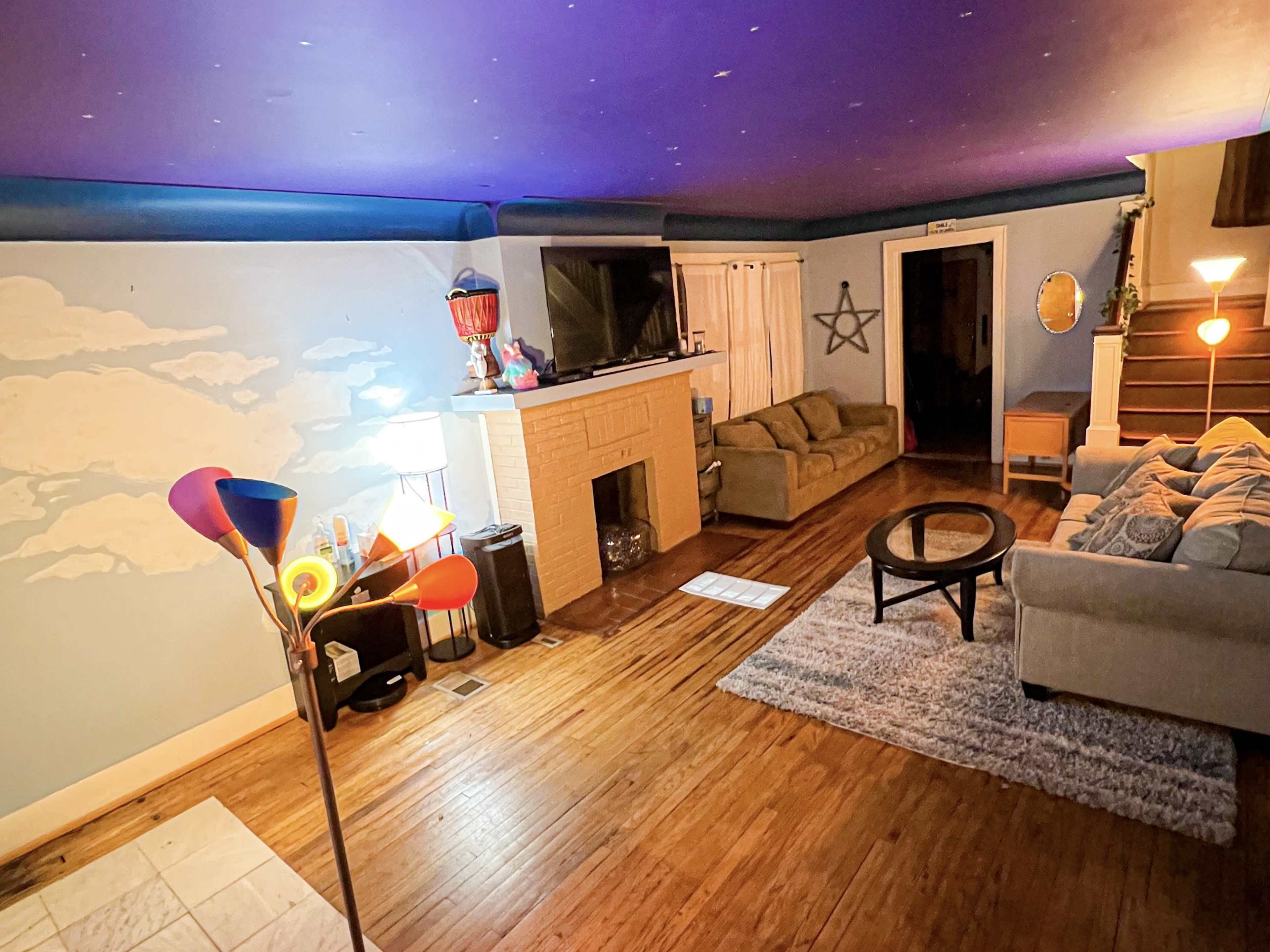 A living room with a purple ceiling, a light-colored wall featuring a cloud mural, a fireplace, two couches, a round coffee table, and various light sources.