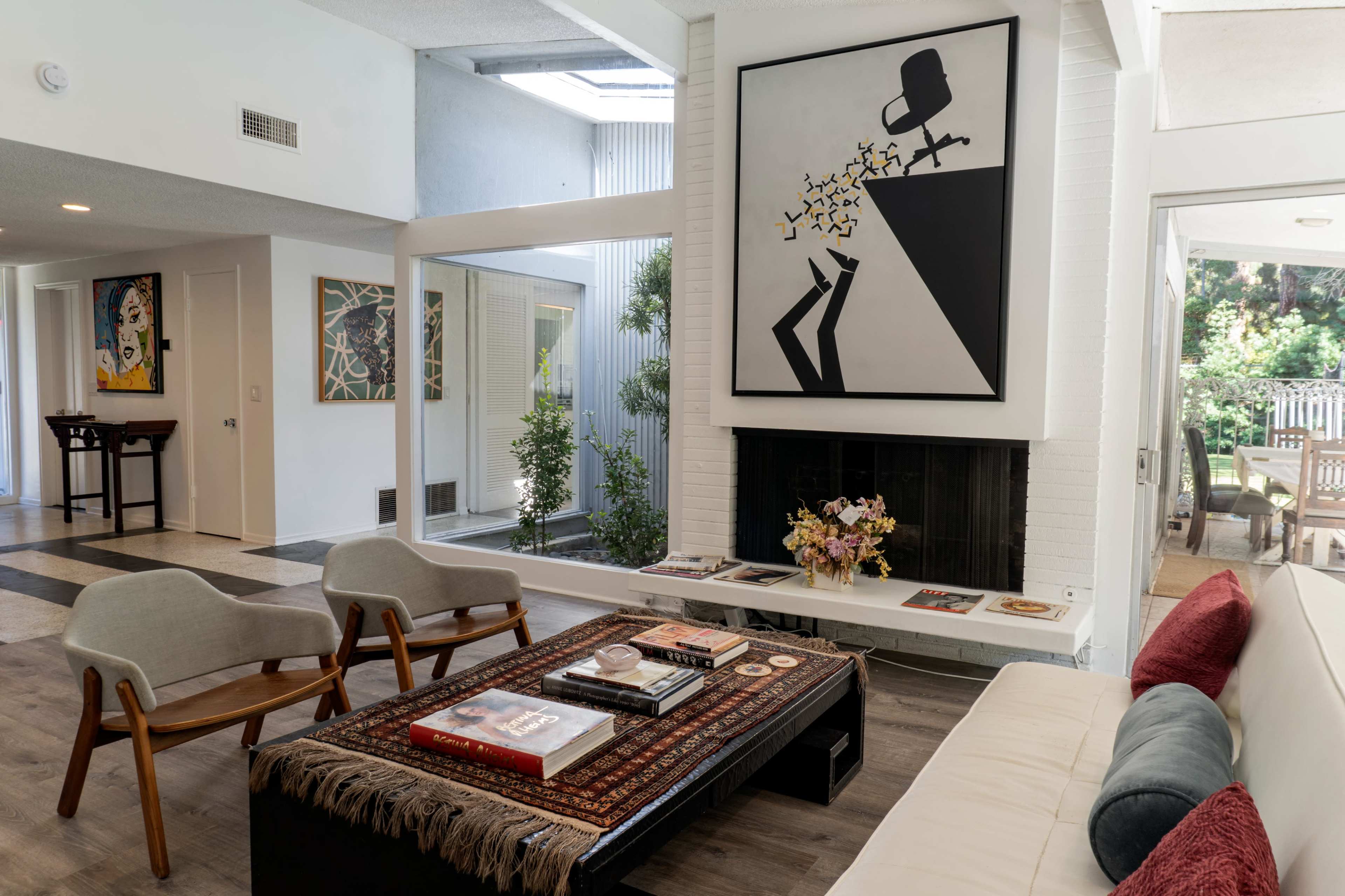 The image shows a modern living room with a large abstract painting above a fireplace, a coffee table with books, and two chairs arranged around it.