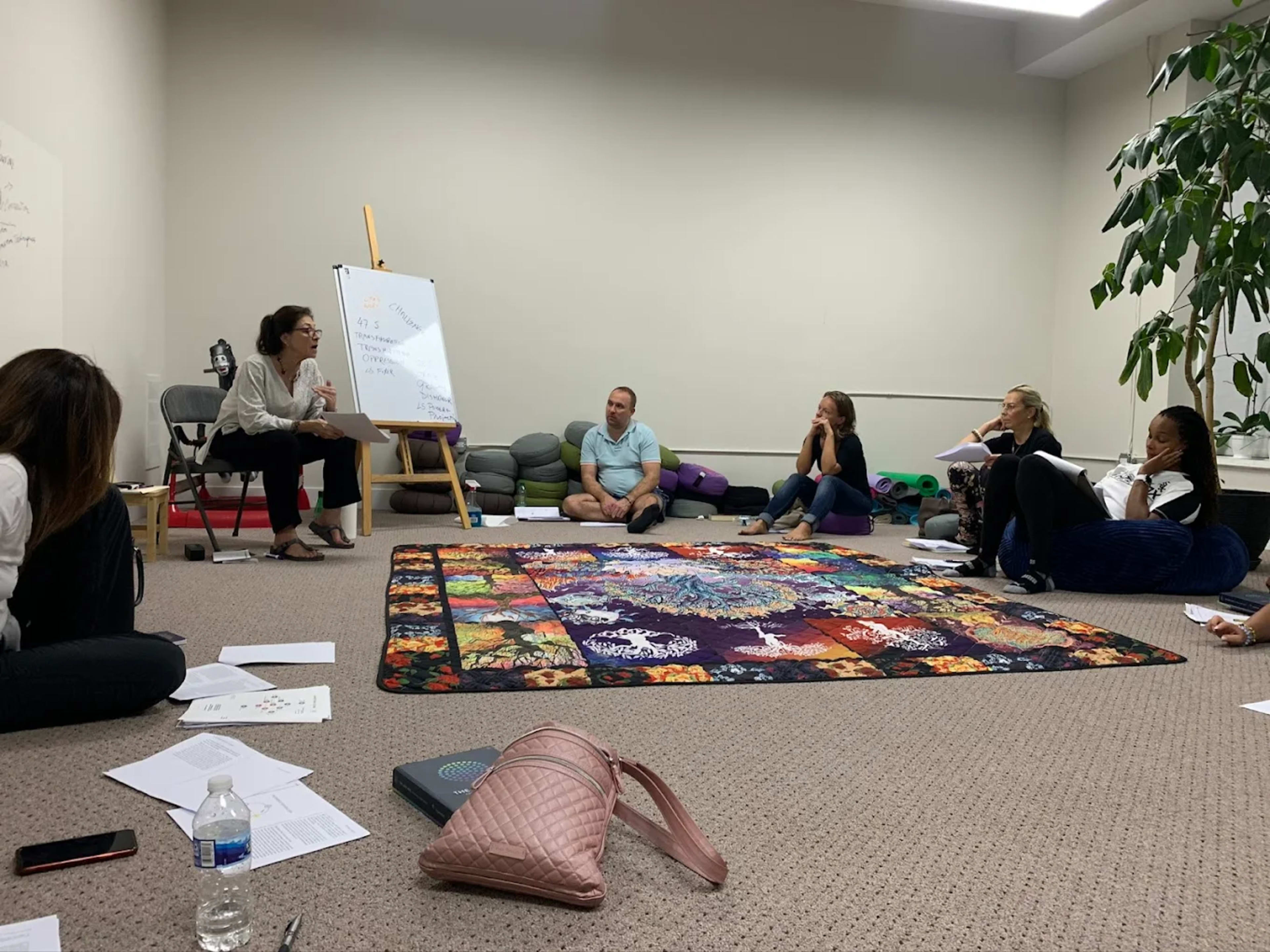 A group of six individuals sits on the floor in a meeting space, engaging in discussion while facing a woman who is speaking next to a whiteboard.