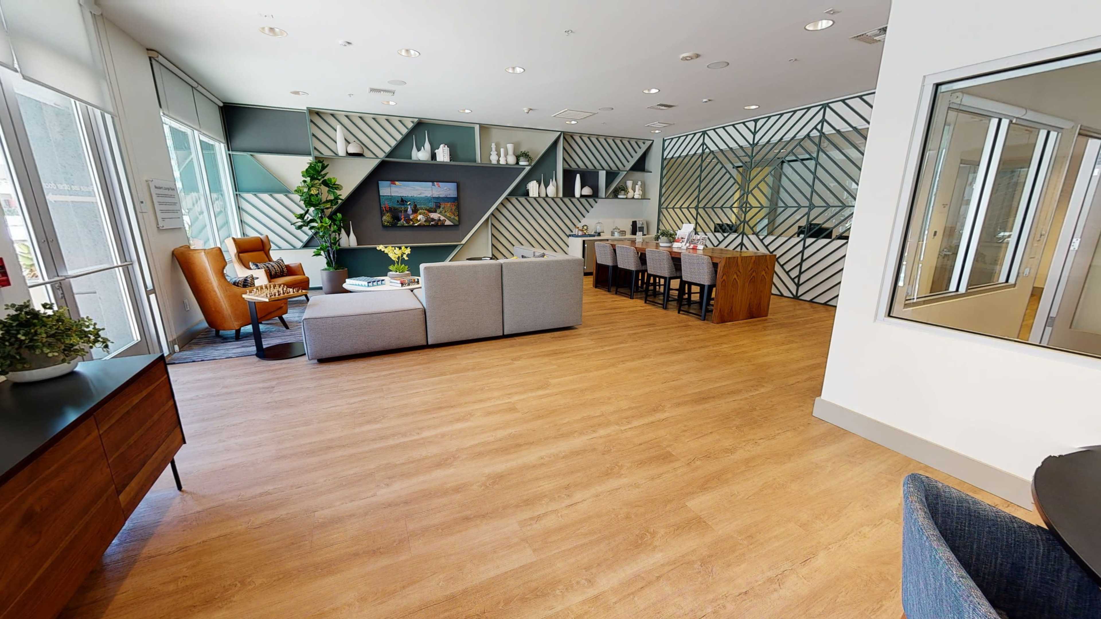 A modern lounge area features a grey sofa, orange armchairs, a television, and a bar-style seating arrangement against a geometric patterned wall.