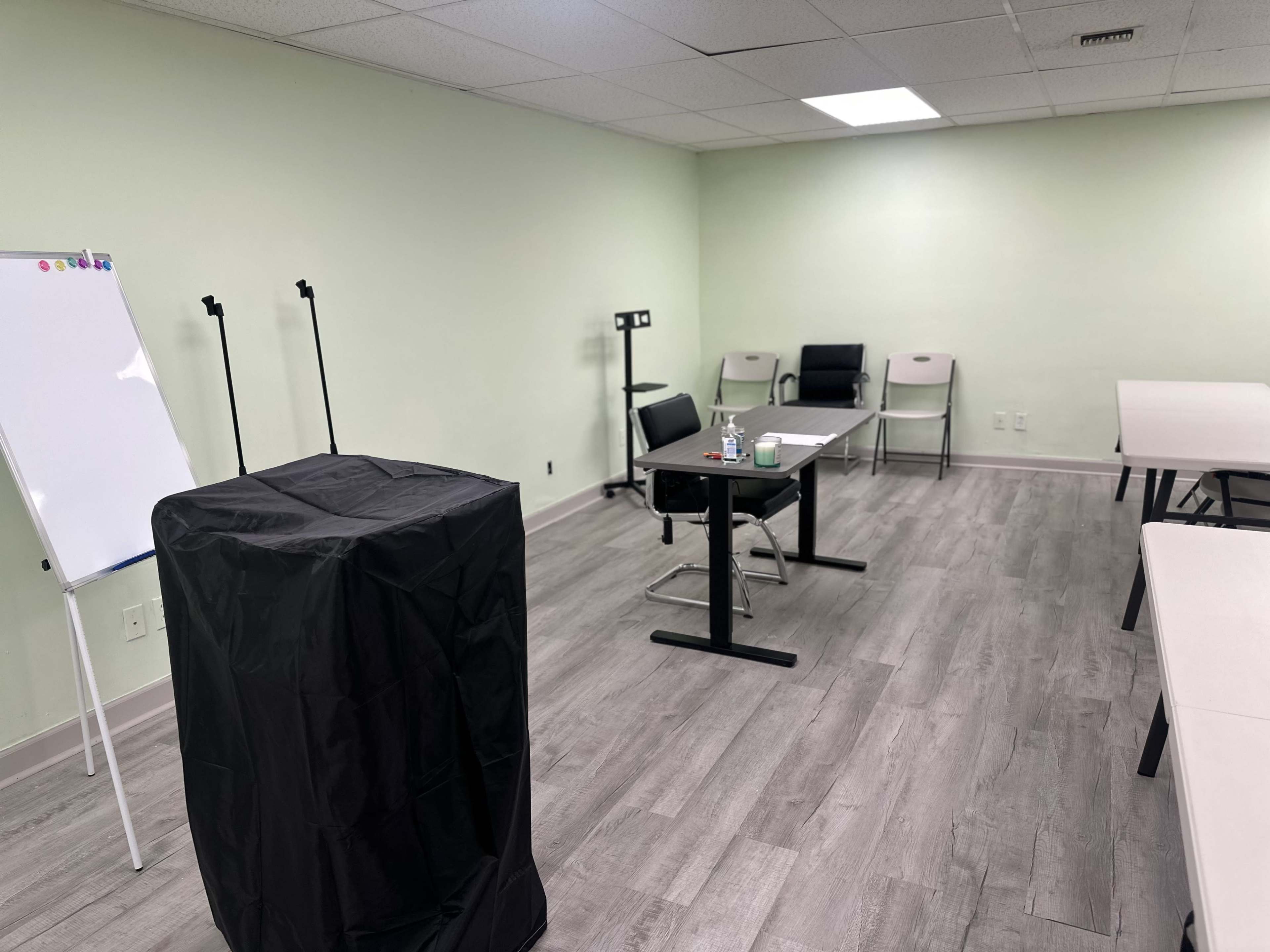 The image shows a sparsely furnished meeting room with a black podium, a desk with a chair, and several empty tables and chairs against light green walls.