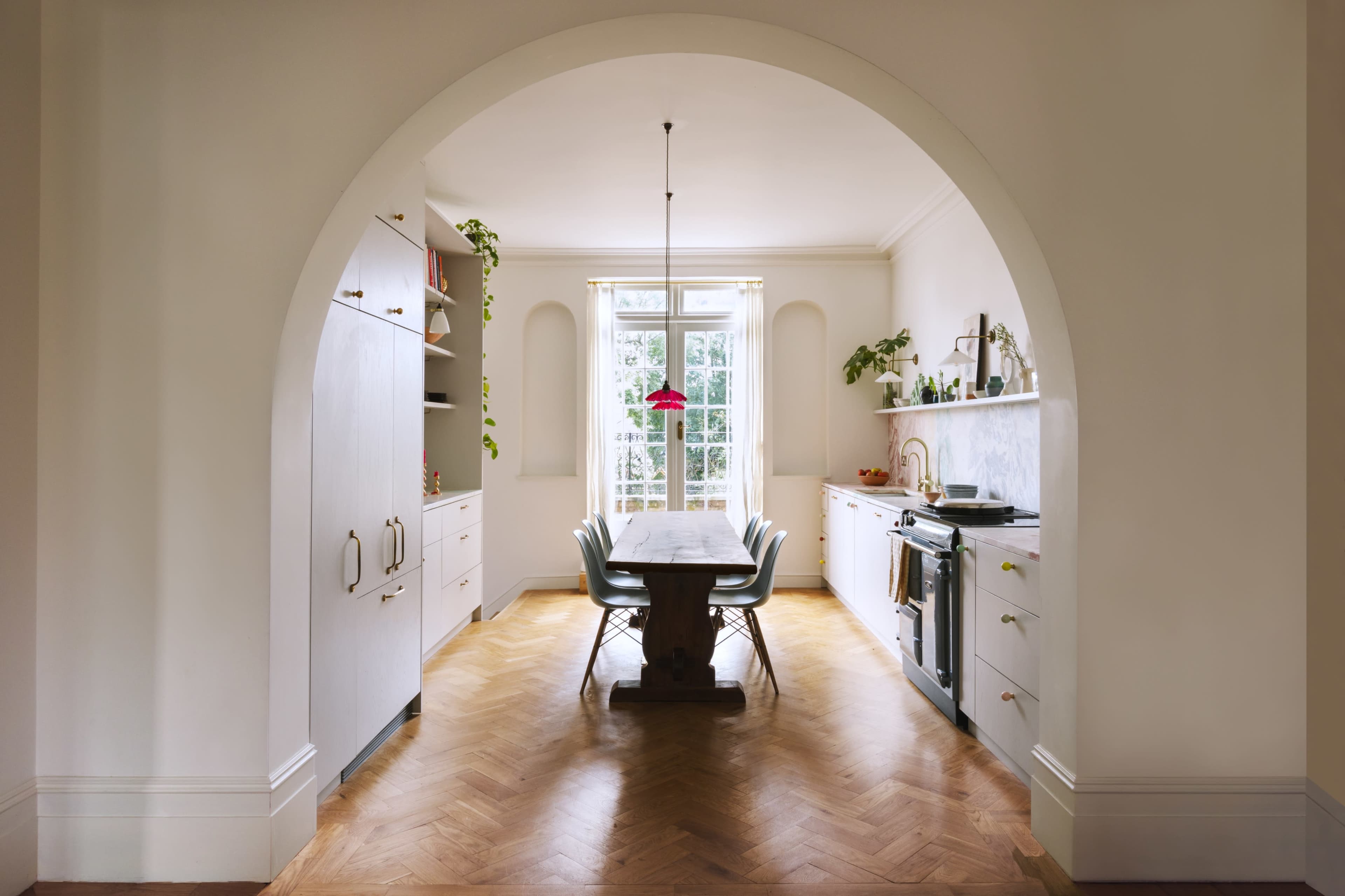 A bright kitchen features a wooden dining table surrounded by chairs, with large windows allowing natural light to fill the space.