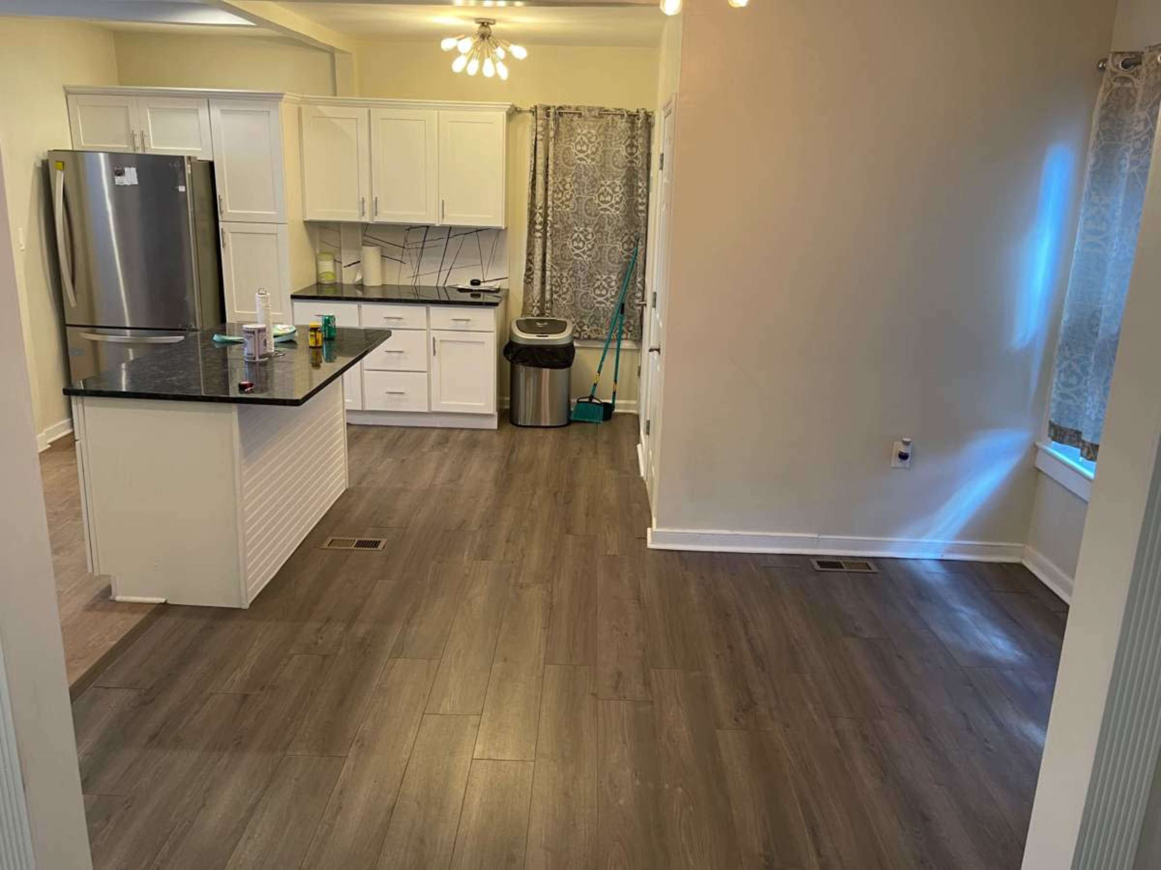 The image shows a bright kitchen with white cabinets, a black countertop, and a spacious area featuring grey laminate flooring.