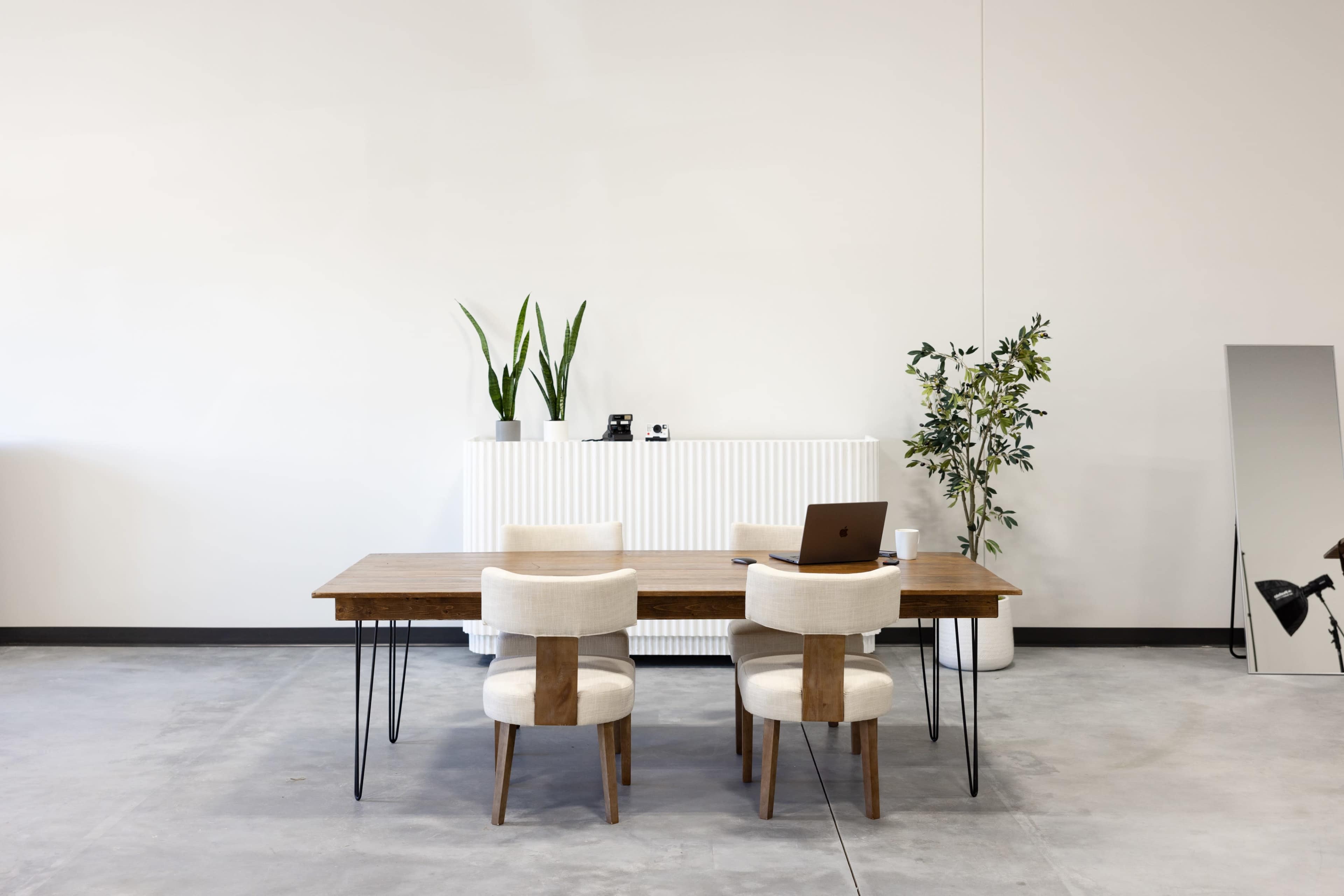 A minimalist office space features a wooden table with three upholstered chairs, a laptop on the table, and plants on a white radiator behind it.