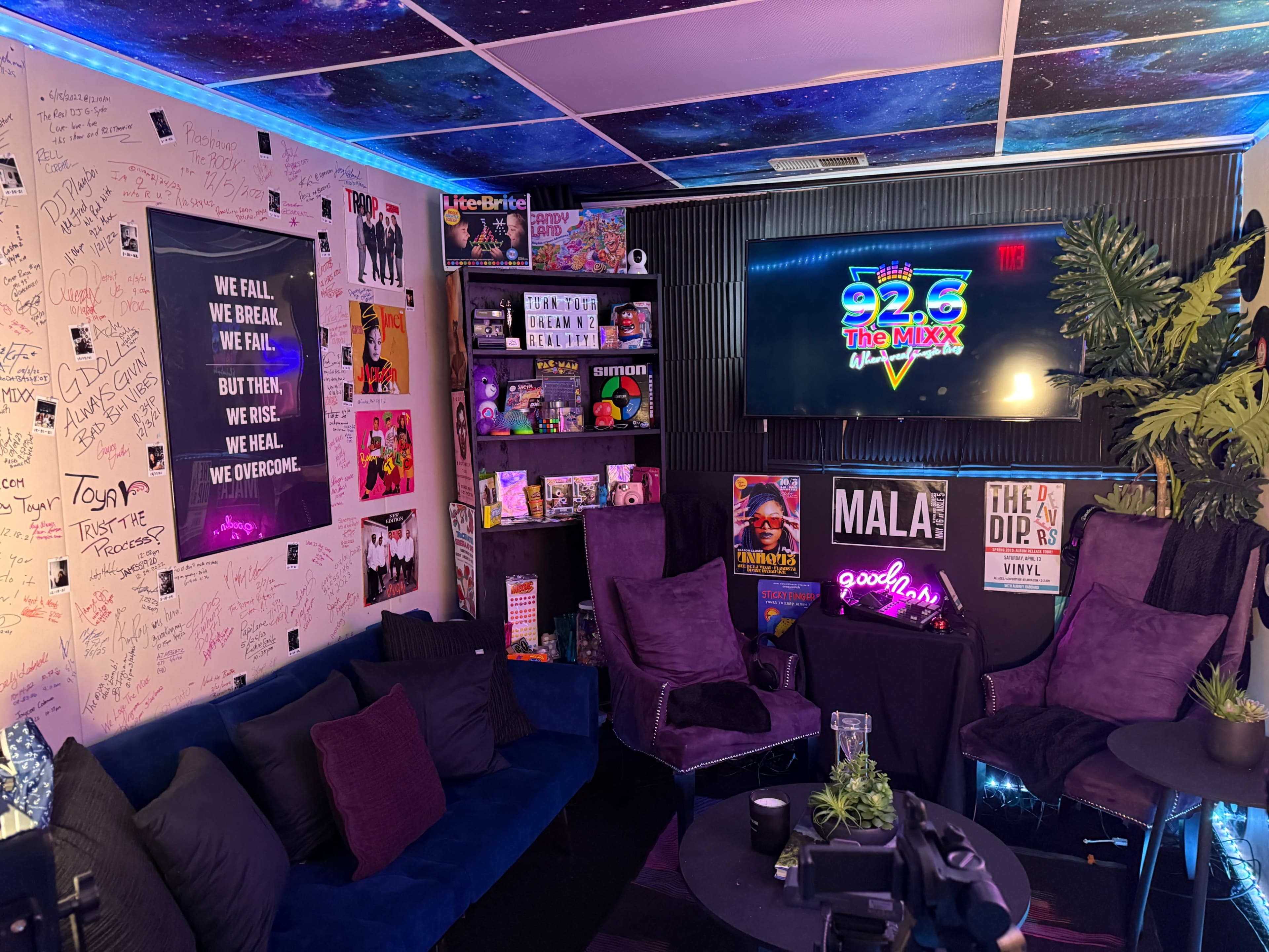 The image shows a vibrant media room decorated with colorful posters, plush seating, and a neon sign that reads "92.6 The Mixx," along with handwritten messages on the walls.