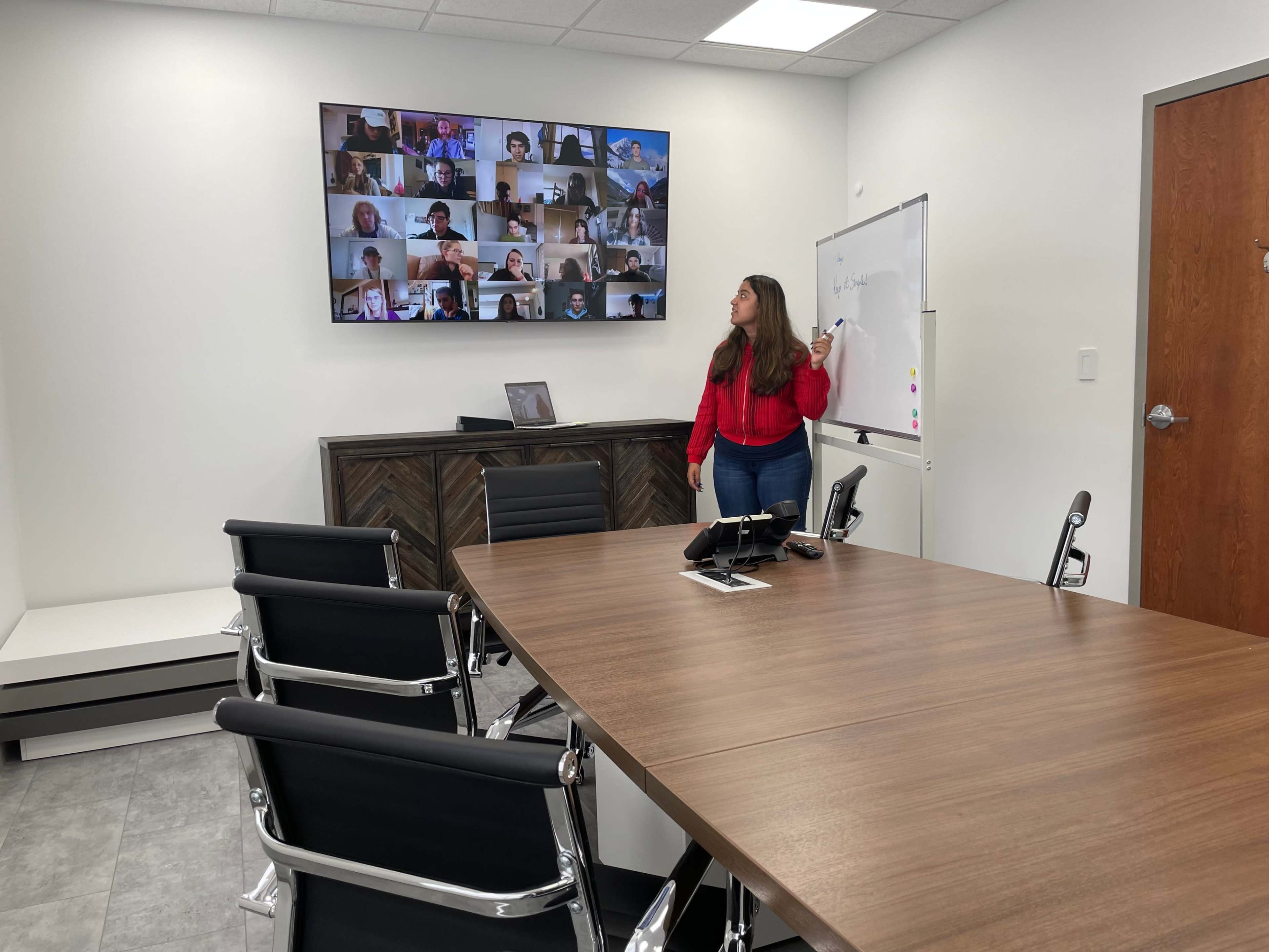A woman stands near a conference table, pointing at a whiteboard while a large screen displays a video conference with multiple participants.