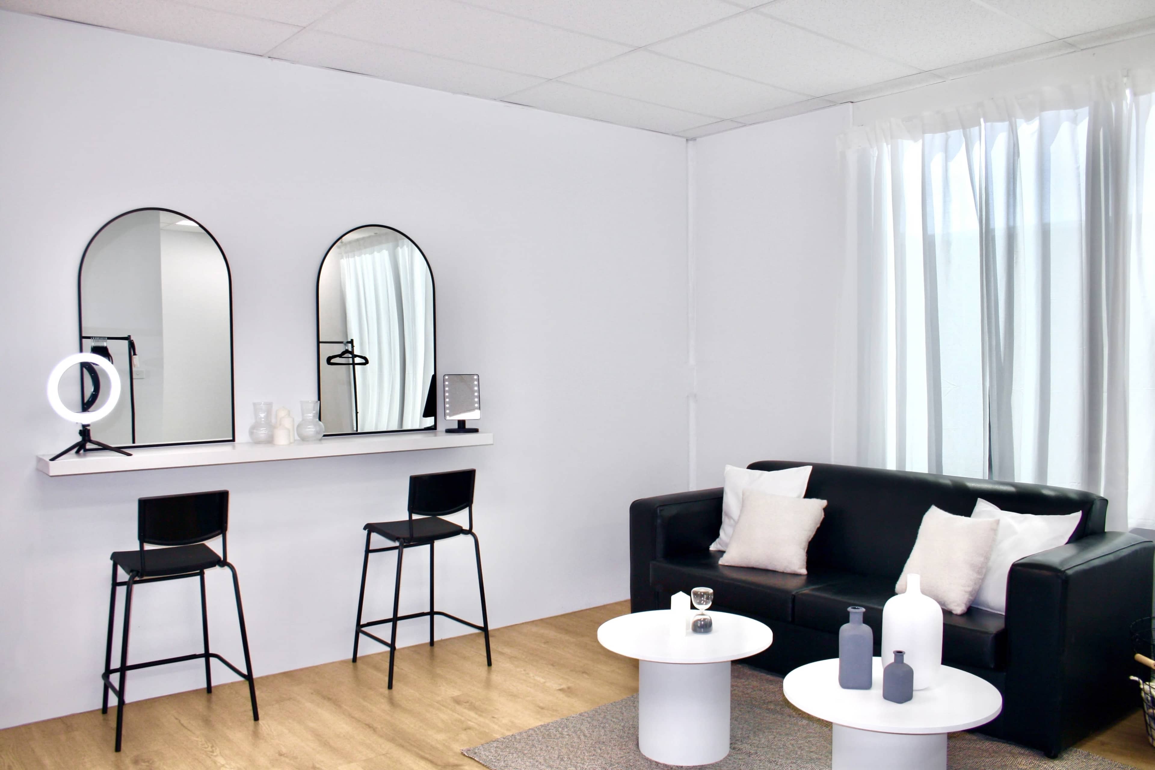 A modern living space featuring a black sofa, two black bar stools at a wall-mounted shelf with mirrors above, and two round white tables adorned with decorative items.