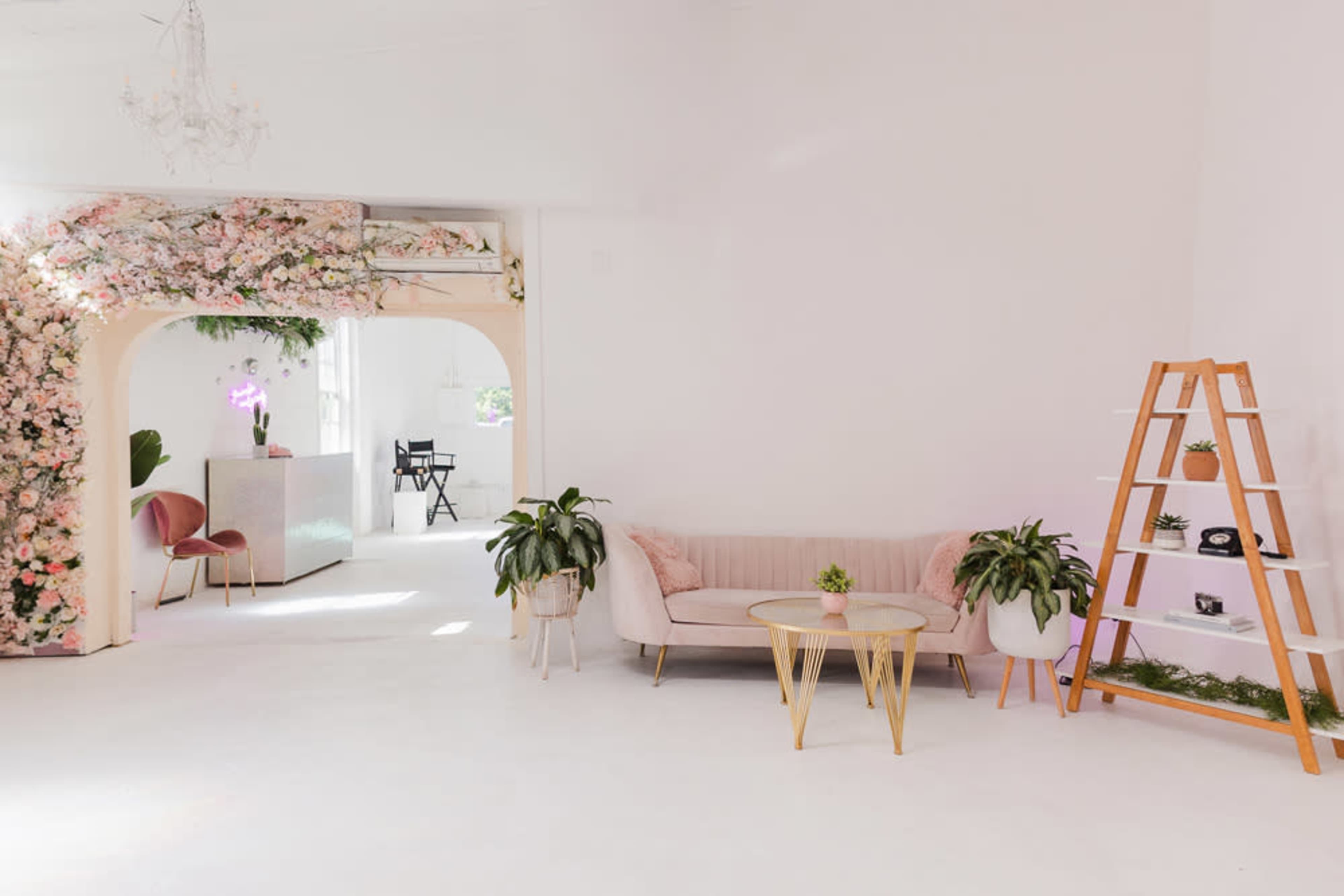 A bright, minimalist room with a pale pink sofa, a wooden ladder shelf, and floral decor around an archway.