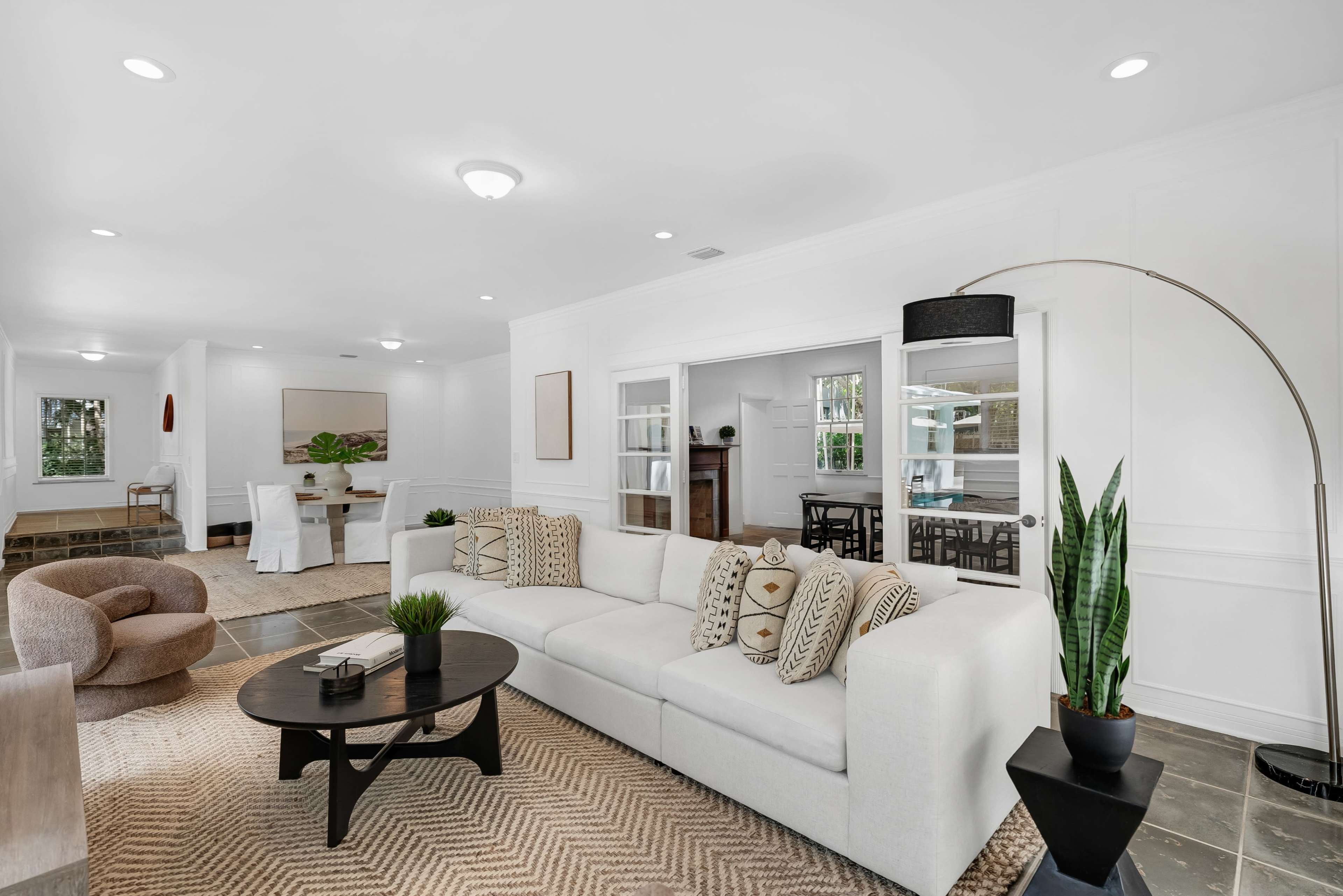 A spacious living area features a white sectional sofa, a round coffee table, and a large plant, with a dining area visible in the background.